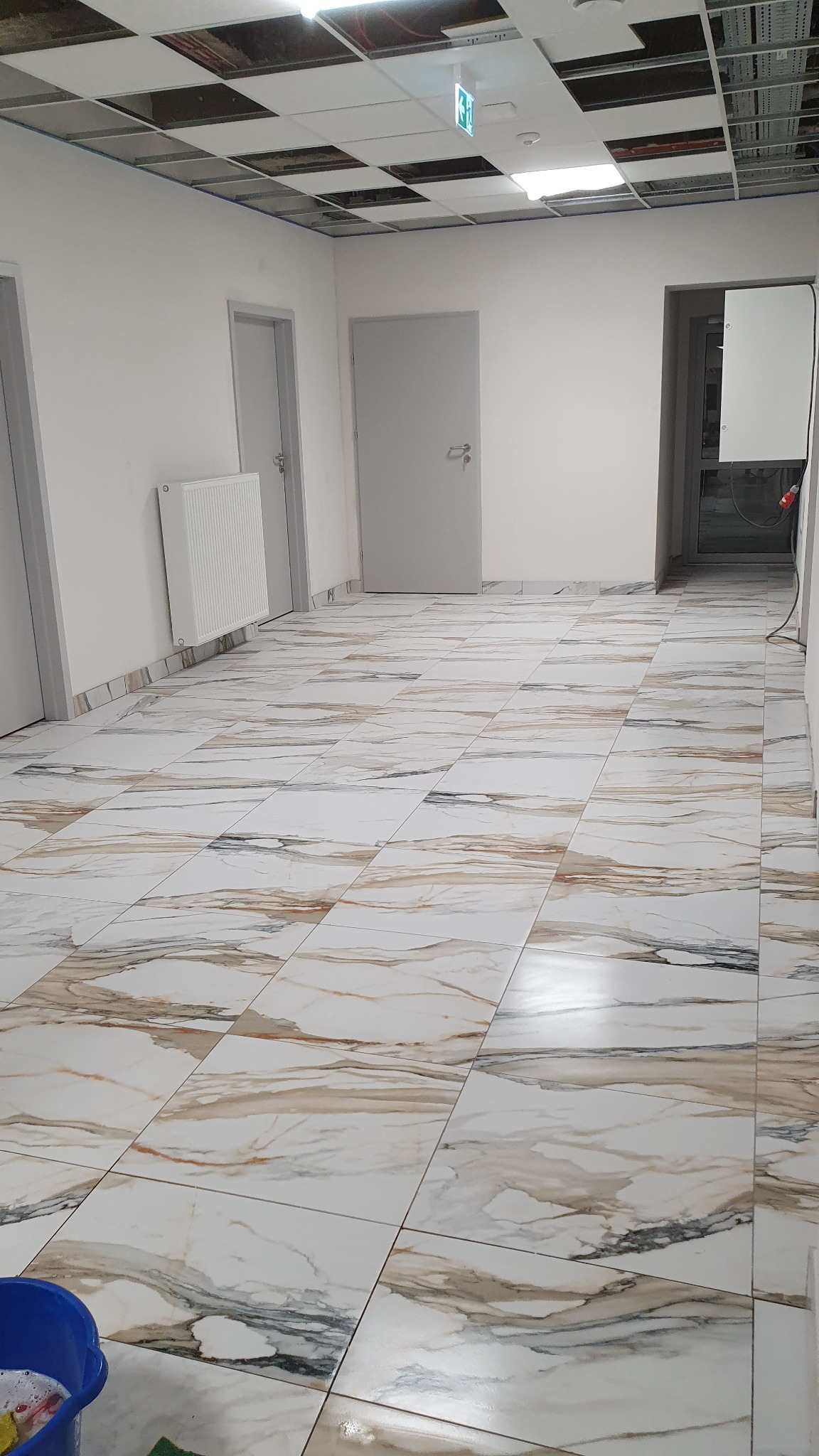 Wnętrze biurowe z marmurową podłogą po umyciu, widoczny fragment sufitu podwieszanego i niebieskie wiadro z mopem w rogu.