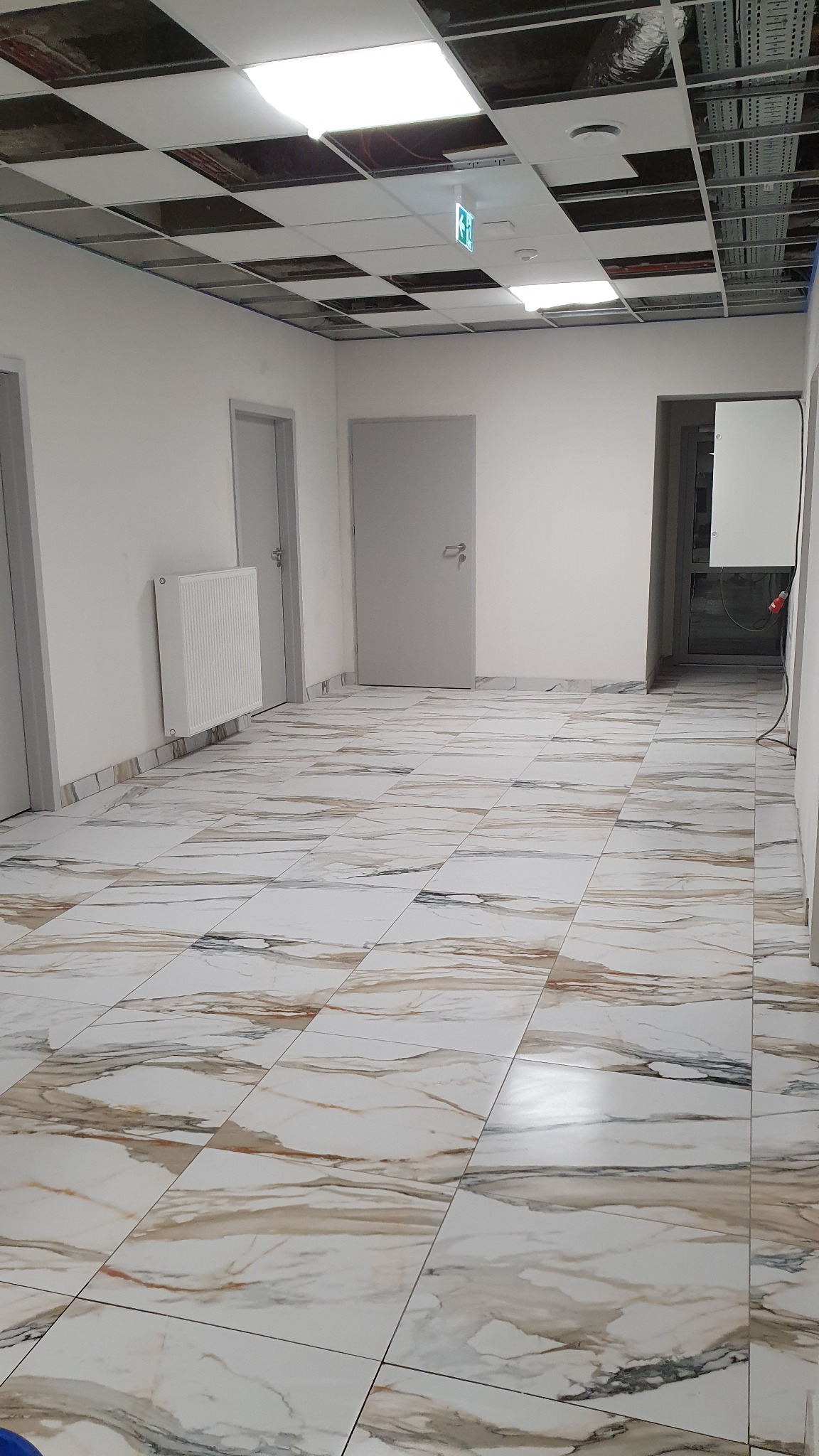 Wnętrze biura z marmurową podłogą, białymi ścianami, grzejnikiem i trzema parami drzwi. Sufit podwieszany z widoczną instalacją i lampami. Na suficie widoczny znak ewakuacyjny.