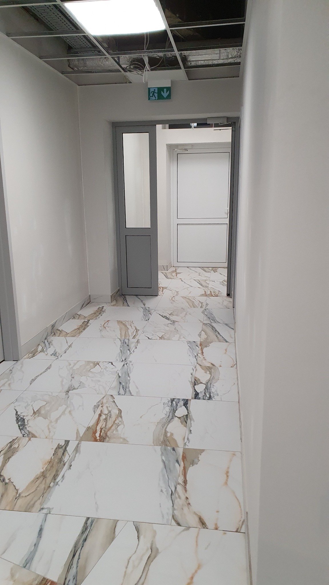 Korytarz biurowy z marmurową podłogą, szarą ościeżnicą i widocznym sufitem podwieszanym z instalacjami.