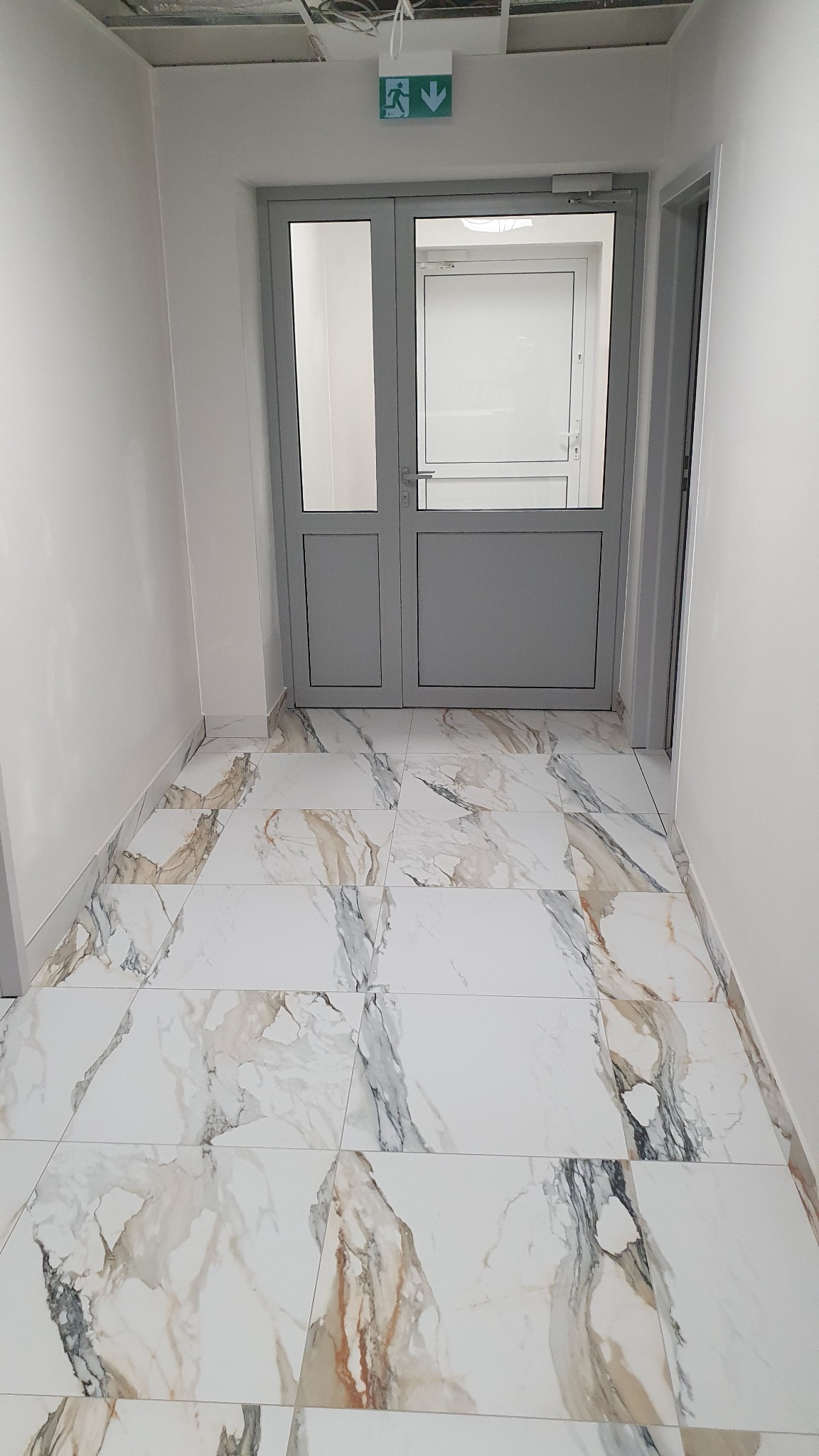 Korytarz z szarymi drzwiami ewakuacyjnymi i podłogą wyłożoną białymi płytkami imitującymi marmur z szarymi i brązowymi żyłami, widoczne przewody pod sufitem.