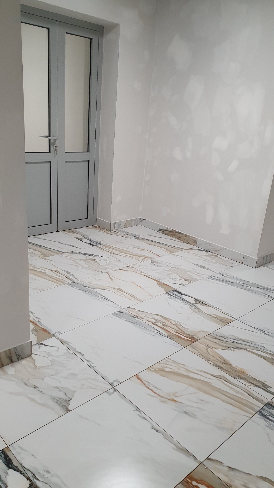 Wnętrze po remoncie: białe ściany z widocznymi śladami szpachlowania, podłoga wyłożona płytkami imitującymi marmur, szare aluminiowe drzwi.