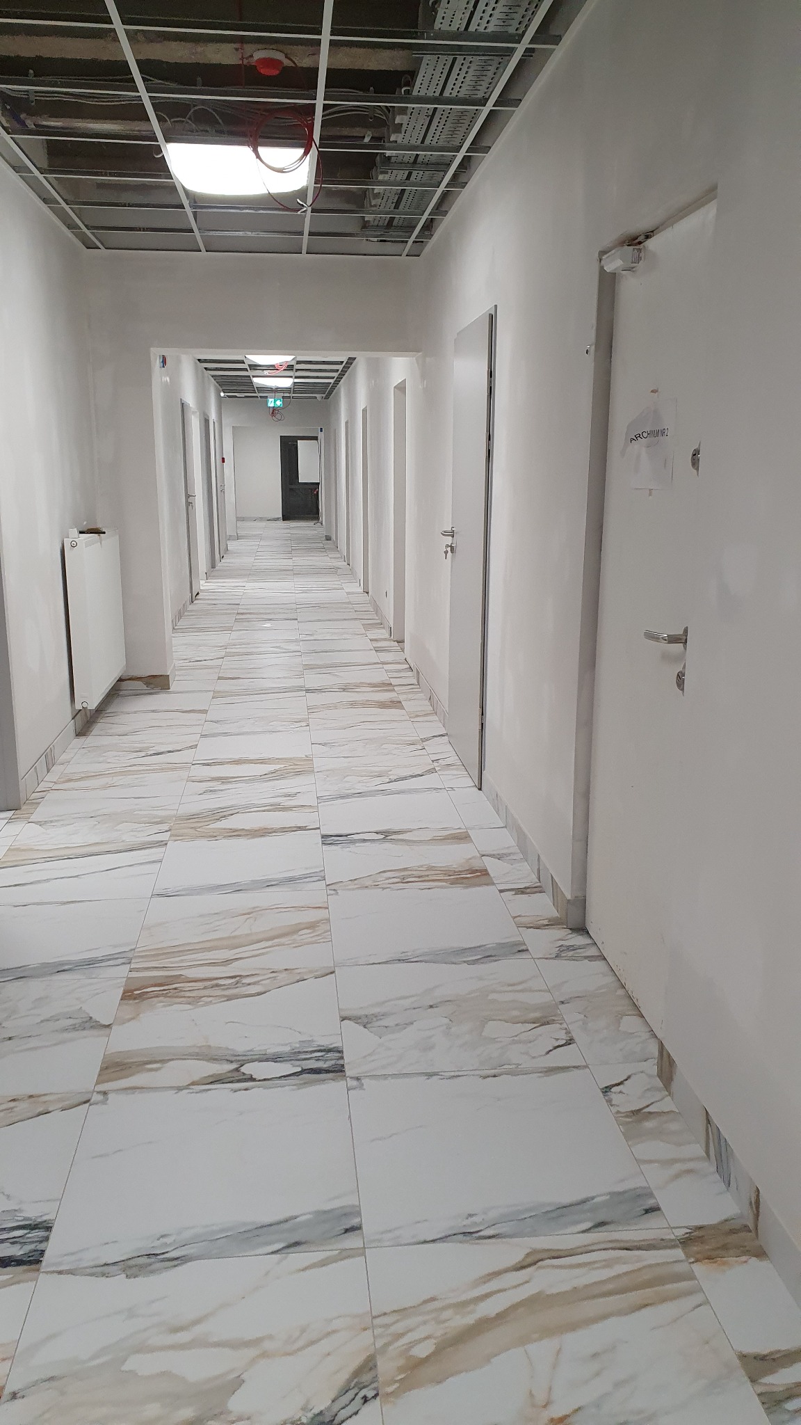 Długi korytarz biurowy w Krakowie po remoncie, z nowymi marmurkowymi płytkami na podłodze i świeżo pomalowanymi ścianami, widok na sufit z odsłoniętymi instalacjami.