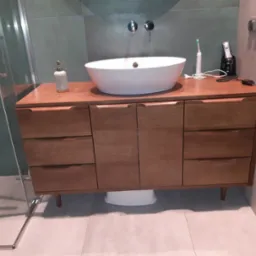 Nowoczesna łazienka z drewnianą szafką pod umywalkę nablatową, widoczna kabina prysznicowa po lewej stronie i płytki w odcieniach szarości na ścianach.