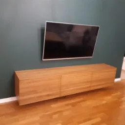 Nowoczesna szafka RTV z jasnego drewna, zawieszona pod telewizorem na tle ściany w kolorze butelkowej zieleni, widoczna podłoga z paneli.