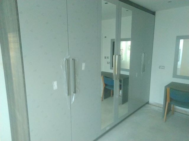 Biała szafa z lustrzanymi drzwiami, częściowo zabezpieczona folią ochronną, w pokoju z biurkiem i krzesłem widocznymi w odbiciu lustra.