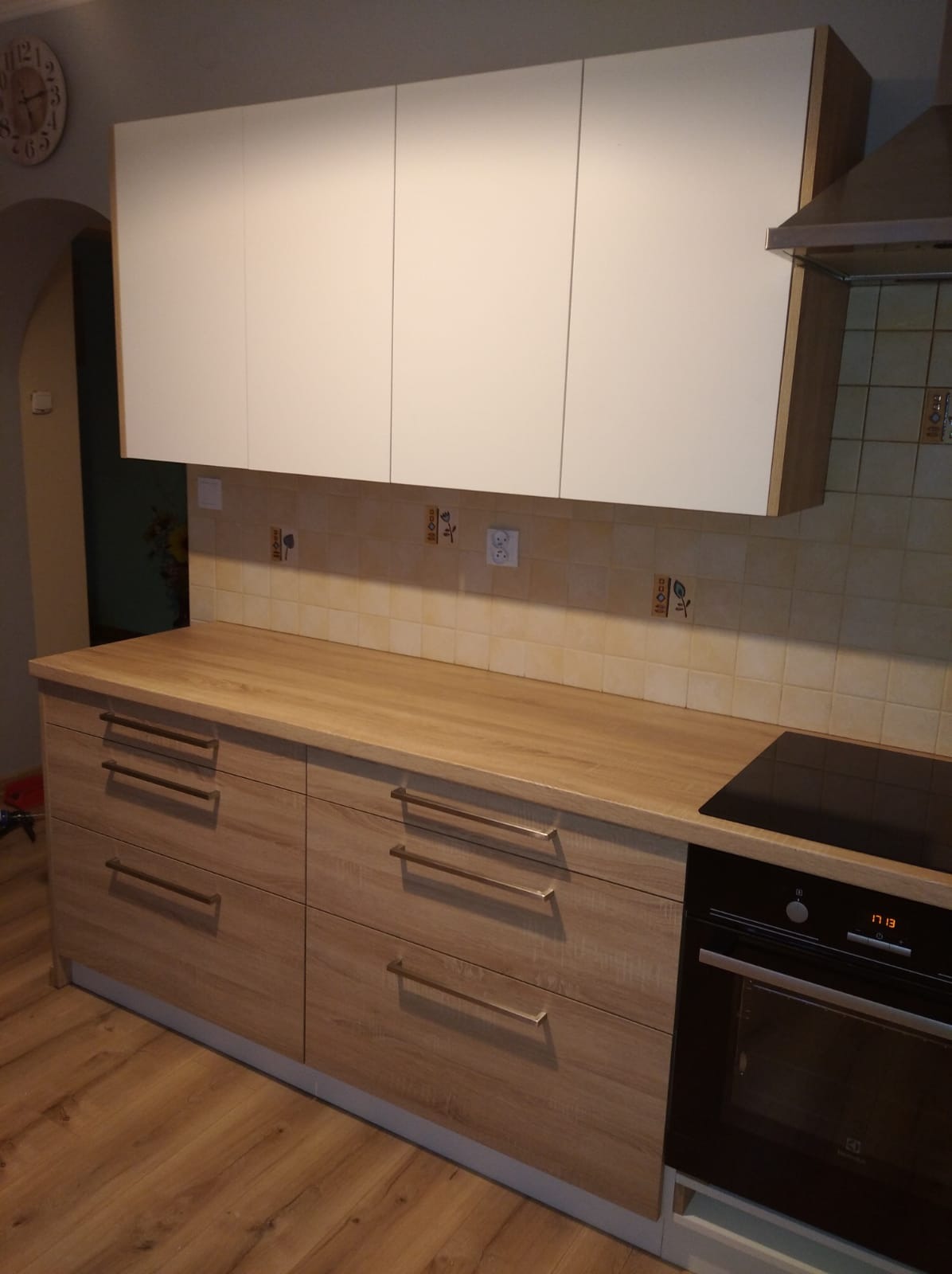 Nowoczesna zabudowa kuchenna z drewnianymi frontami szafek i białymi szafkami wiszącymi, płyta indukcyjna wkomponowana w blat, widoczny fragment piekarnika i okapu.