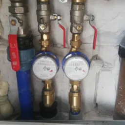 Zestaw dwóch wodomierzy z zaworami odcinającymi zamontowany na ścianie z widocznym filtrem mechanicznym. Instalacja wykonana z mosiądzu i rur z tworzywa sztucznego.