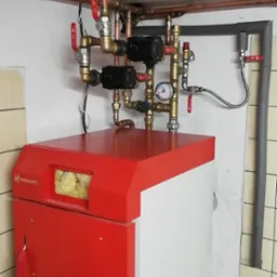 Czerwono-biały piec centralnego ogrzewania z widocznymi rurami miedzianymi, zaworami i manometrem, zamontowany w pomieszczeniu wyłożonym kafelkami.