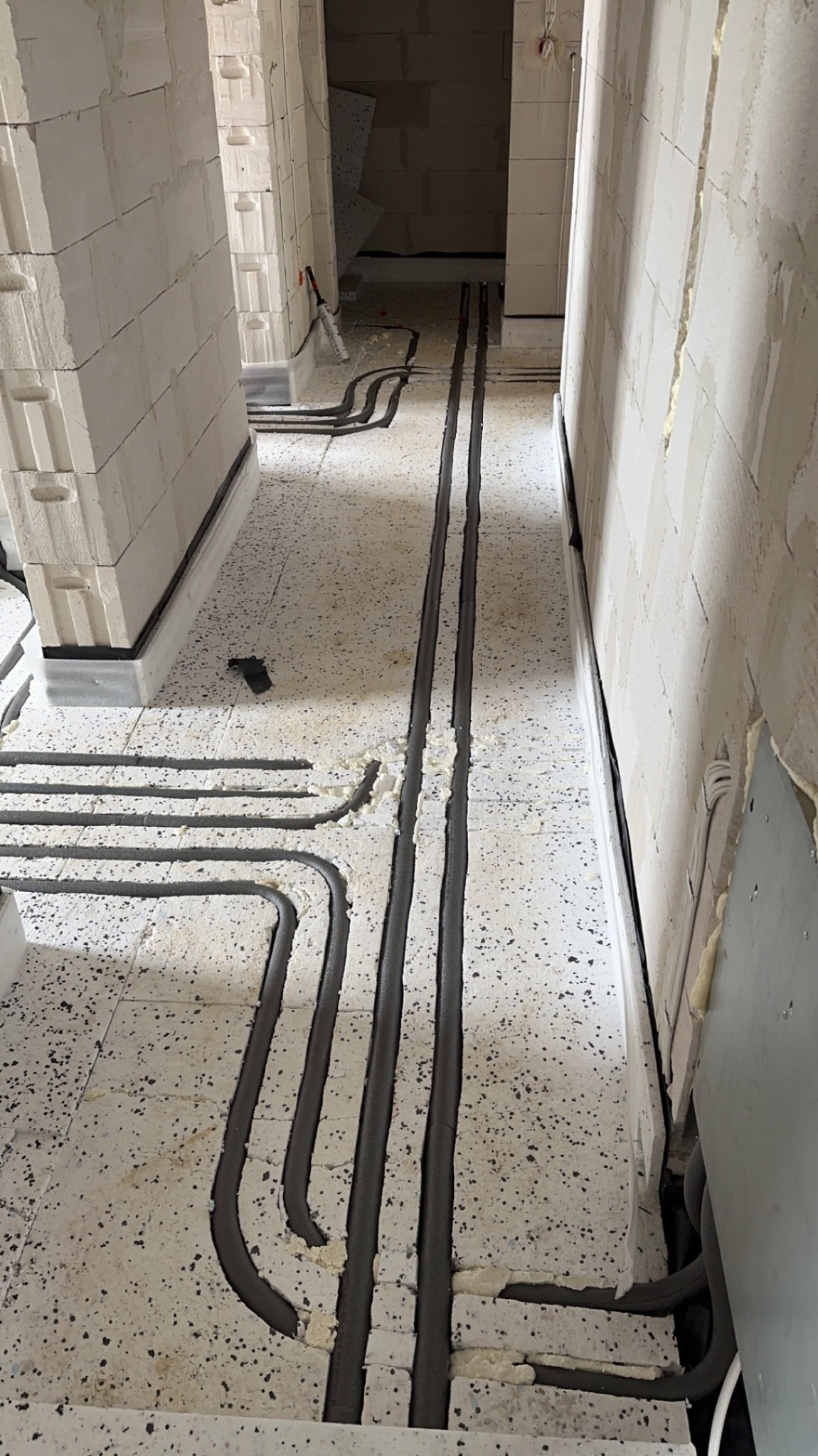 Rury instalacji wodnej ułożone w wycięciach w styropianie pod podłogą, zabezpieczone pianką montażową, widok w surowym stanie budynku z białymi ścianami z bloczków.