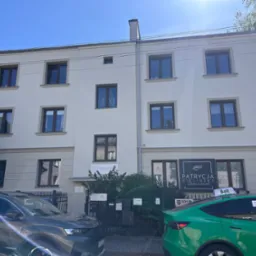 Odnowiona elewacja dwupiętrowego budynku z brązowymi oknami, widoczne szyldy salonów kosmetycznych i zaparkowane samochody. Słoneczny dzień.