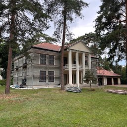 ORCO Sp. z o.o. - Dwupiętrowy dom z kolumnami w trakcie prac elewacyjnych, otoczony rusztowaniami i drzewami. Widoczne materiały budowlane na trawniku.