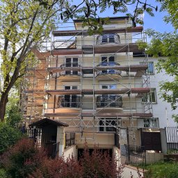 ORCO Sp. z o.o. - Budynek wielorodzinny w trakcie malowania elewacji, otoczony rusztowaniami. Widoczne balkony z ozdobnymi balustradami i zieleń drzew. Słoneczny dzień.