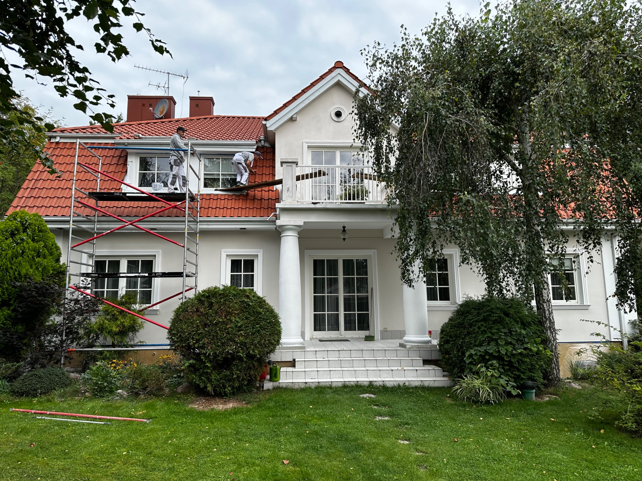 Dwóch malarzy na rusztowaniu przy kremowej elewacji domu z czerwoną dachówką, jeden maluje okap, drugi pracuje przy oknie na poddaszu, widoczny fragment trawnika i drzewo.
