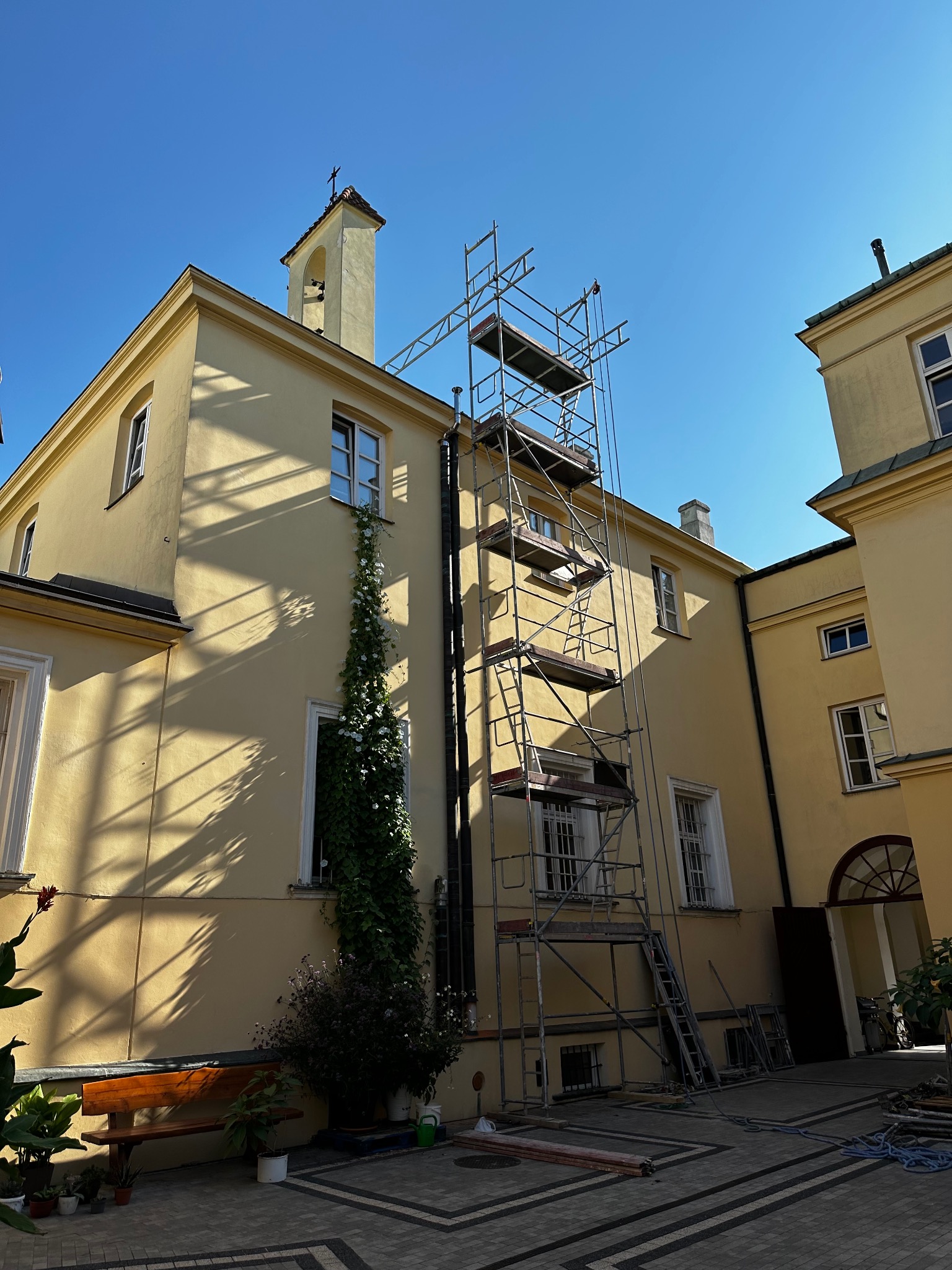 Żółta elewacja budynku z wieżą i dzwonem, częściowo zasłonięta rusztowaniem, z pnącą rośliną przy oknie