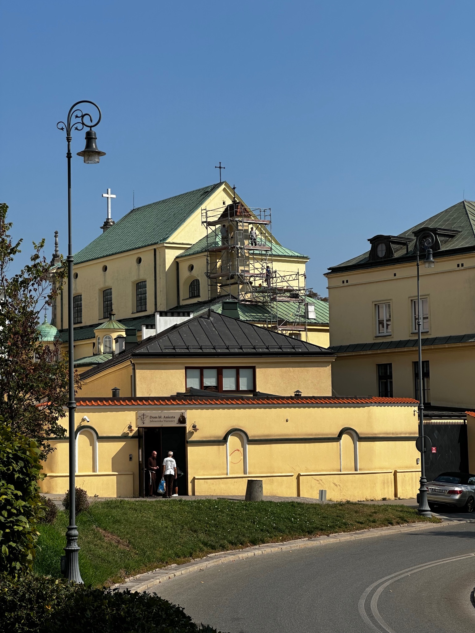 Żółty budynek kościoła z zielonym dachem w trakcie renowacji, widoczne rusztowania, błękitne niebo, latarnia uliczna i fragment ulicy.