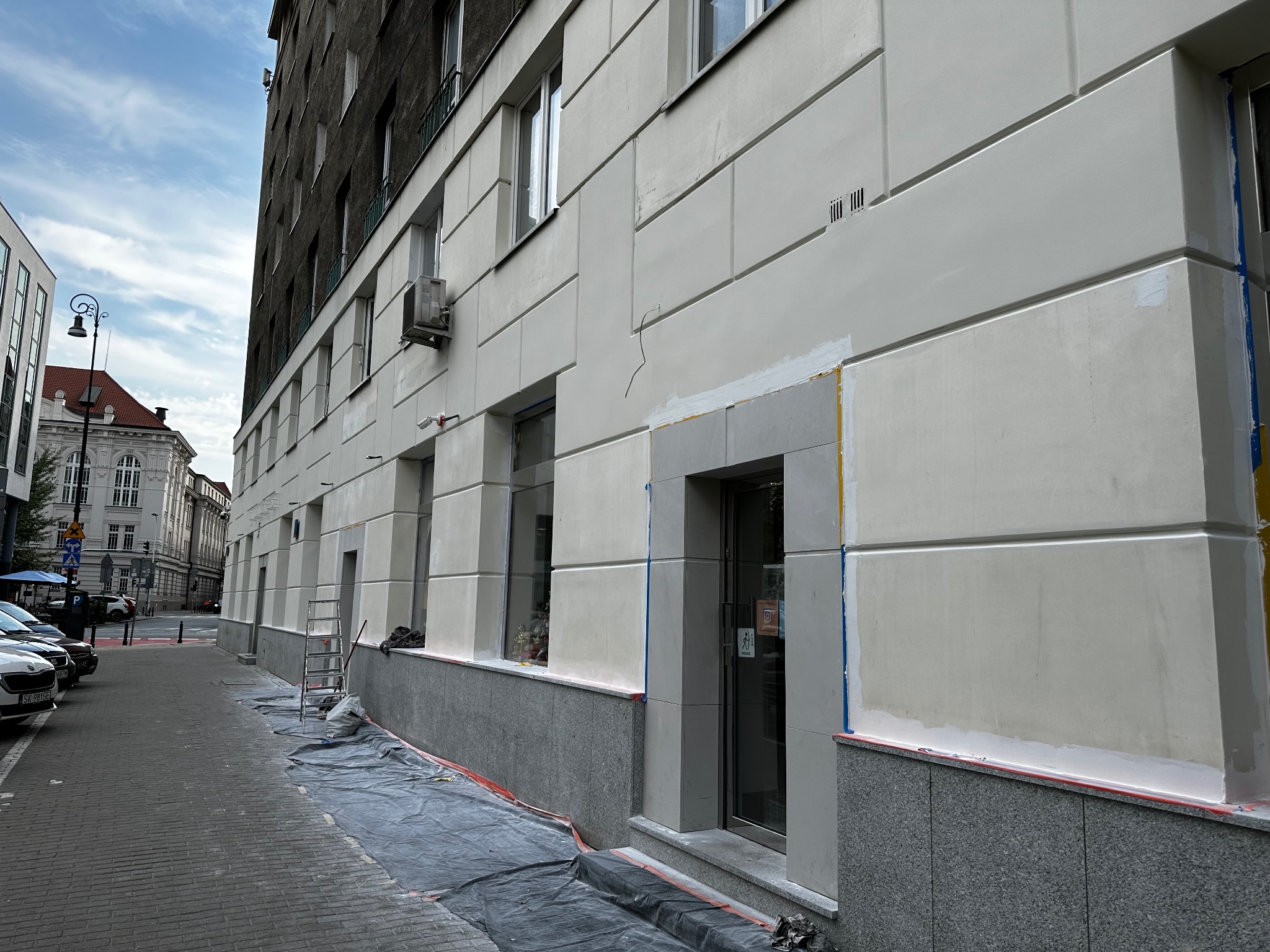 Fragment odnowionej elewacji budynku z widocznymi śladami prac przygotowawczych, zabezpieczona folią i taśmą malarską, z drabiną opartą o ścianę na tle chodnika i ulicy.
