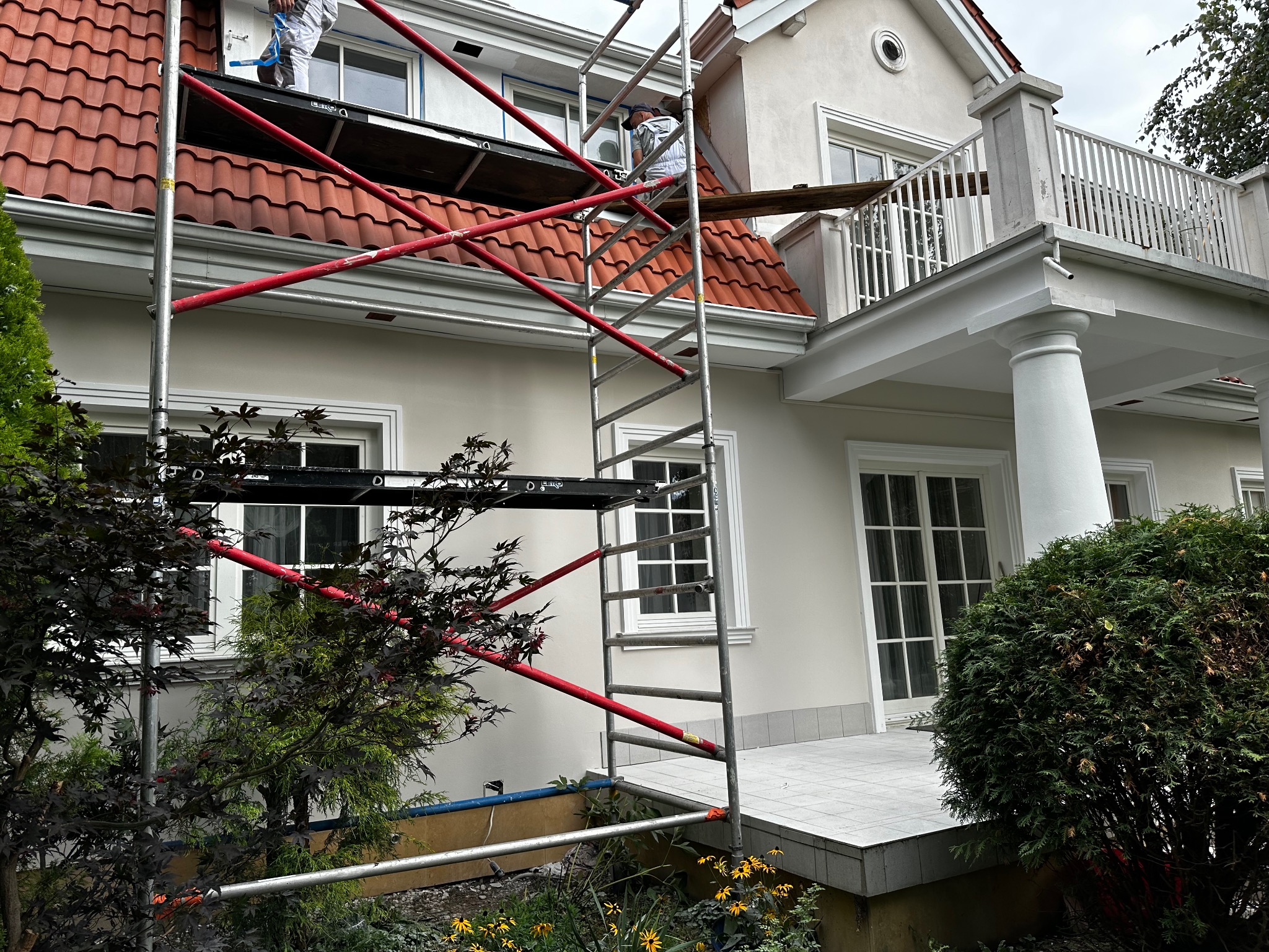 Dwóch pracowników na rusztowaniu maluje elewację budynku z czerwonym dachem, widoczne okna z białymi ramami, białe kolumny i balkon