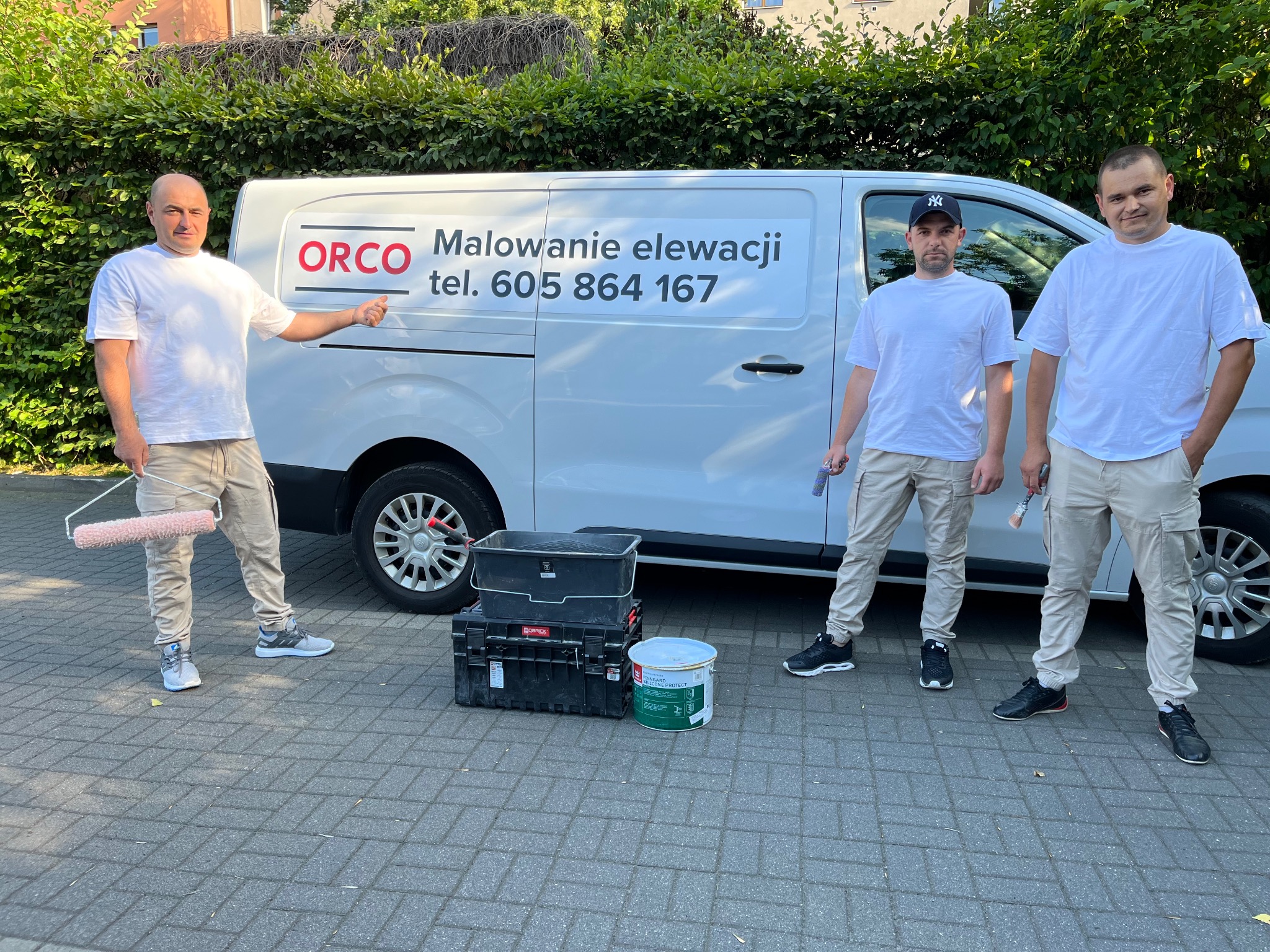 Trzech mężczyzn w roboczych ubraniach stoi przed białym busem z napisem 'ORCO Malowanie elewacji tel. 605 864 167', obok widoczne narzędzia malarskie i puszka farby.
