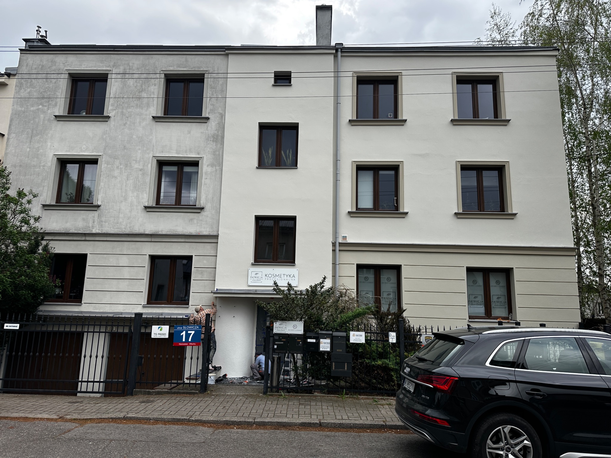 Malowanie elewacji budynku usługowego w trakcie realizacji. Widoczni pracownicy, rusztowanie, narzędzia. Część elewacji pomalowana na biało, druga w szarym kolorze.