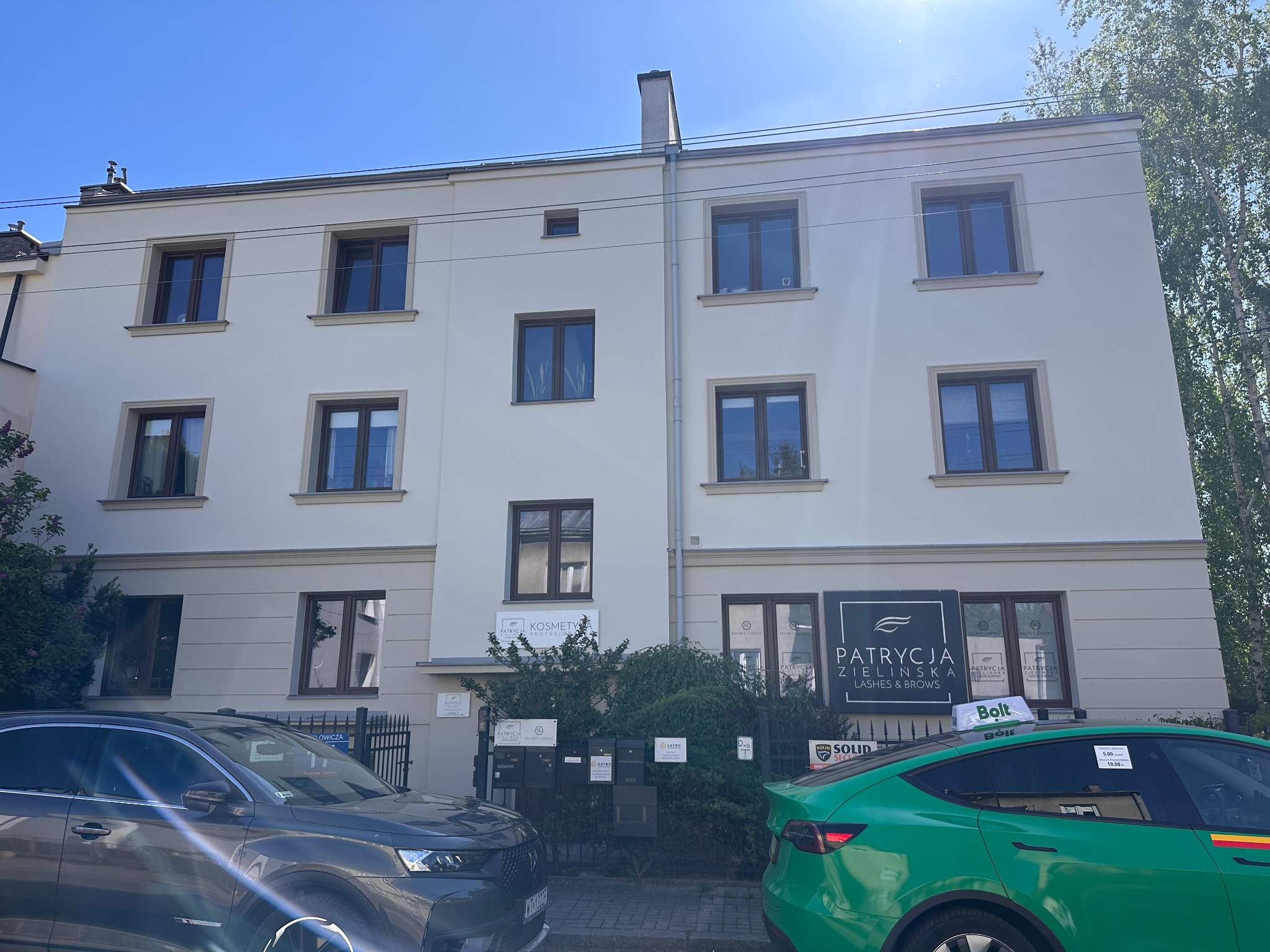 Odnowiona elewacja dwupiętrowego budynku z brązowymi oknami, widoczne szyldy salonów kosmetycznych i zaparkowane samochody. Słoneczny dzień.