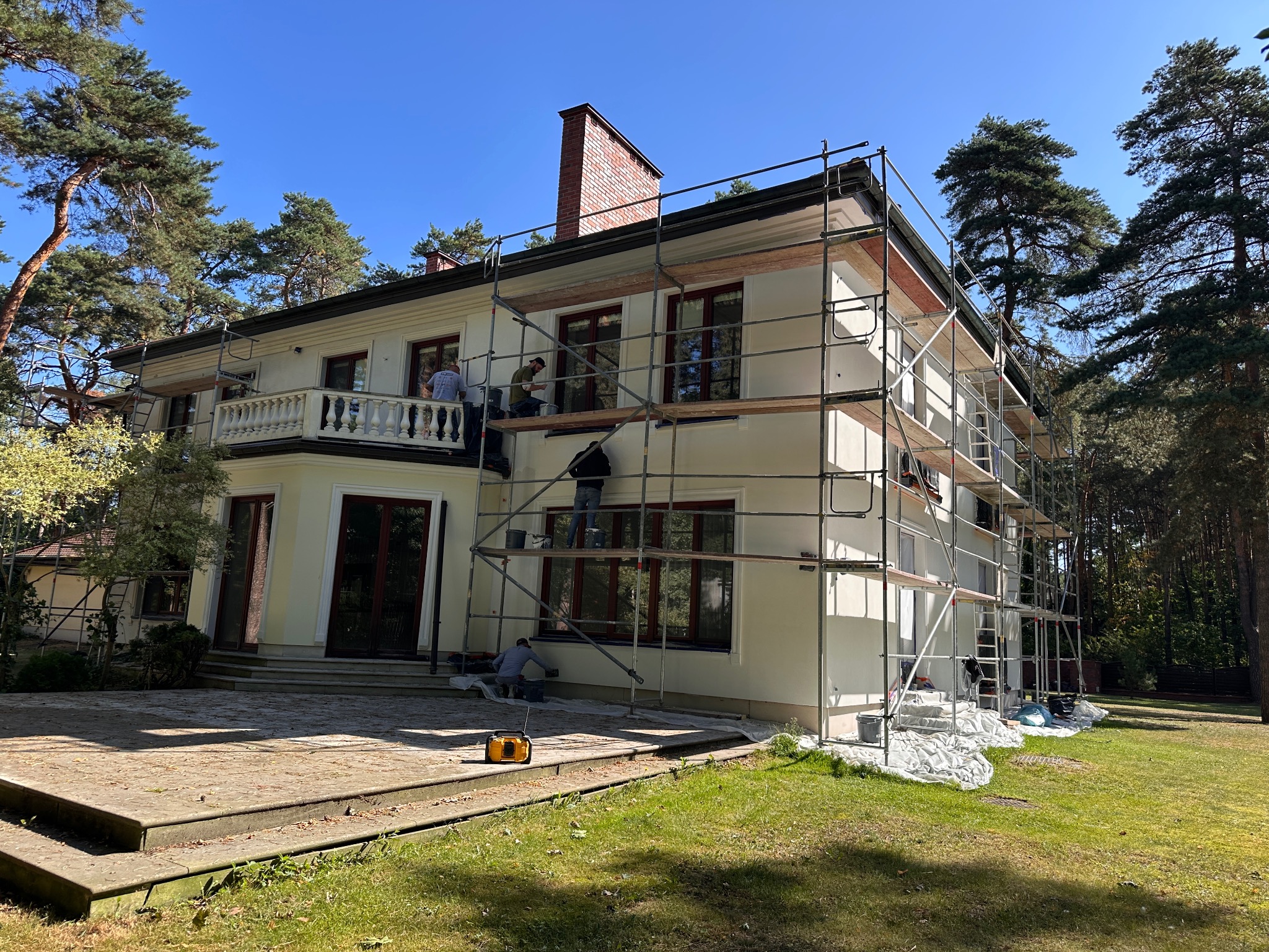 Ekipa maluje elewację dwupiętrowego domu w stylu klasycznym, używając rusztowań. Widoczny fragment ogrodu i błękitne niebo. Prace w toku, częściowo pomalowana ściana.