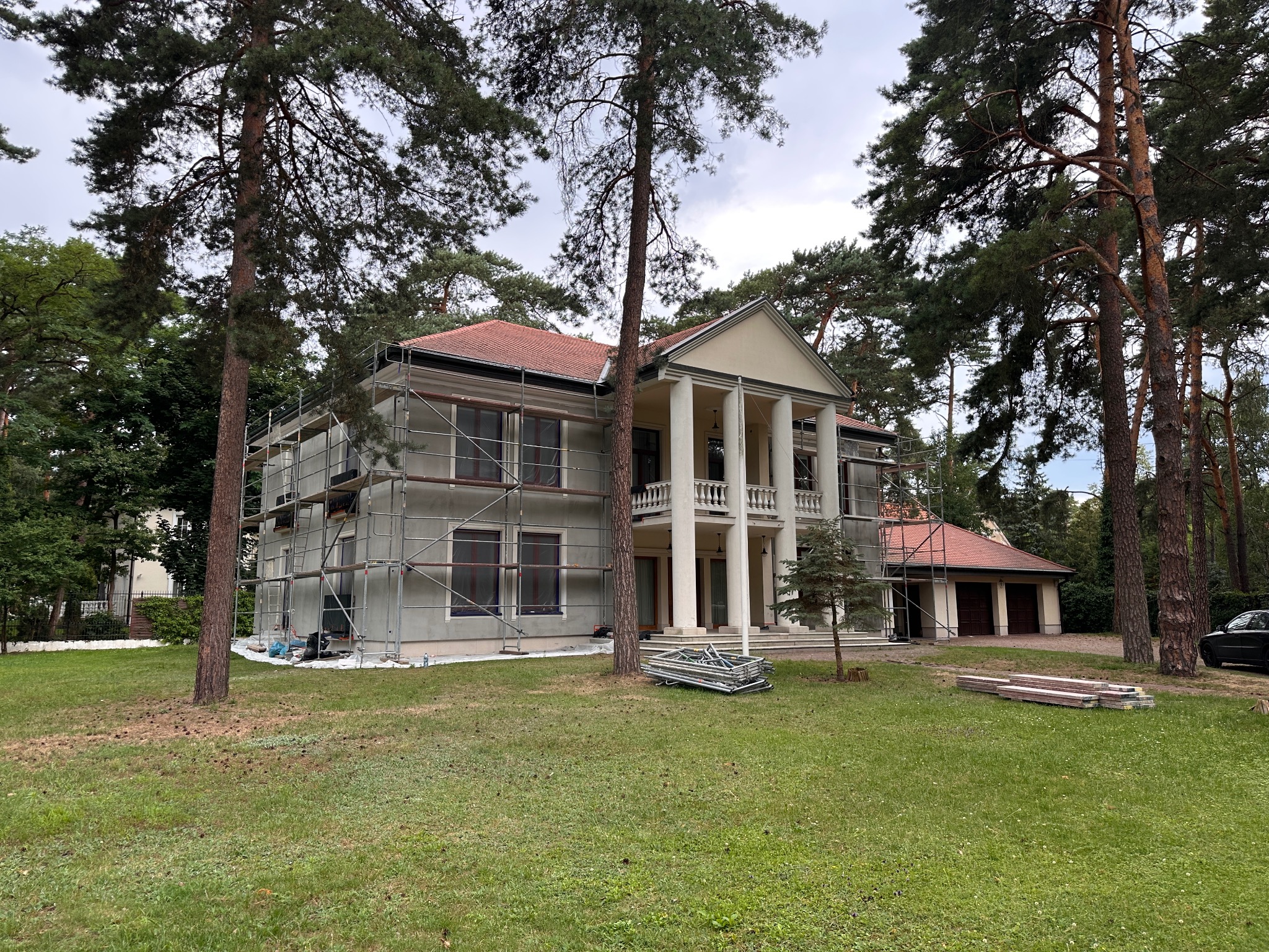 Dwupiętrowy dom z kolumnami w trakcie prac elewacyjnych, otoczony rusztowaniami i drzewami. Widoczne materiały budowlane na trawniku.