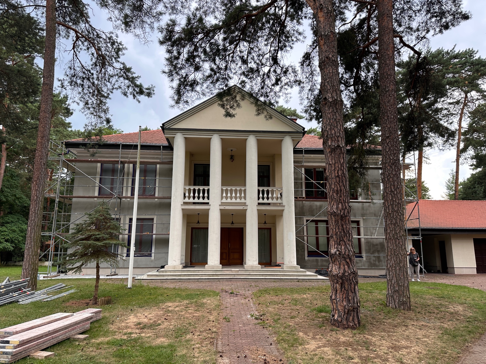 Dwupiętrowy dom z kolumnami w trakcie prac elewacyjnych, widoczne rusztowania i przygotowane deski. Częściowo otynkowana fasada, drzewa na pierwszym planie.