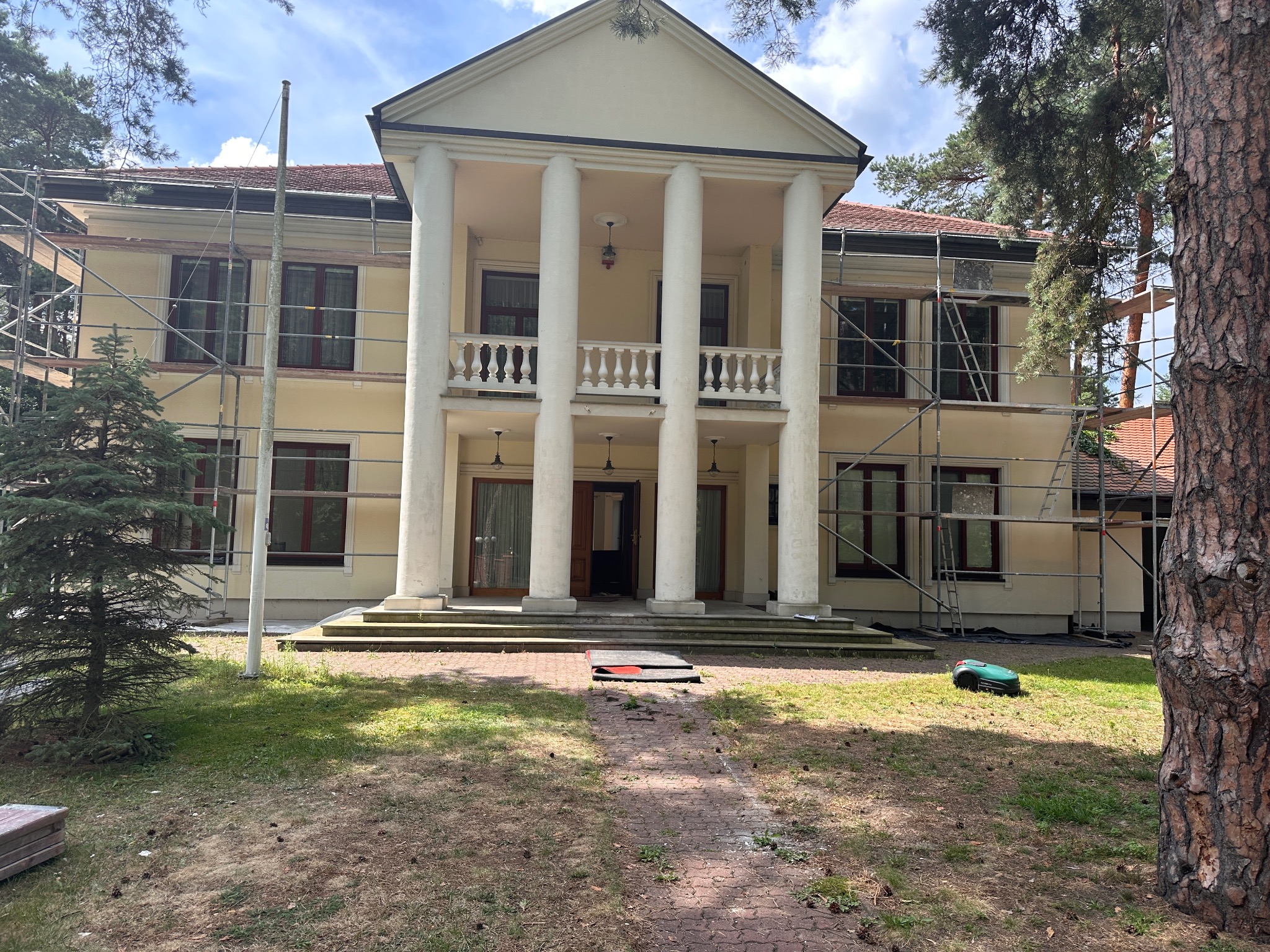 Dwukondygnacyjny budynek z kolumnami w trakcie malowania elewacji, otoczony rusztowaniami i zielenią. Widoczny robot koszący trawę.