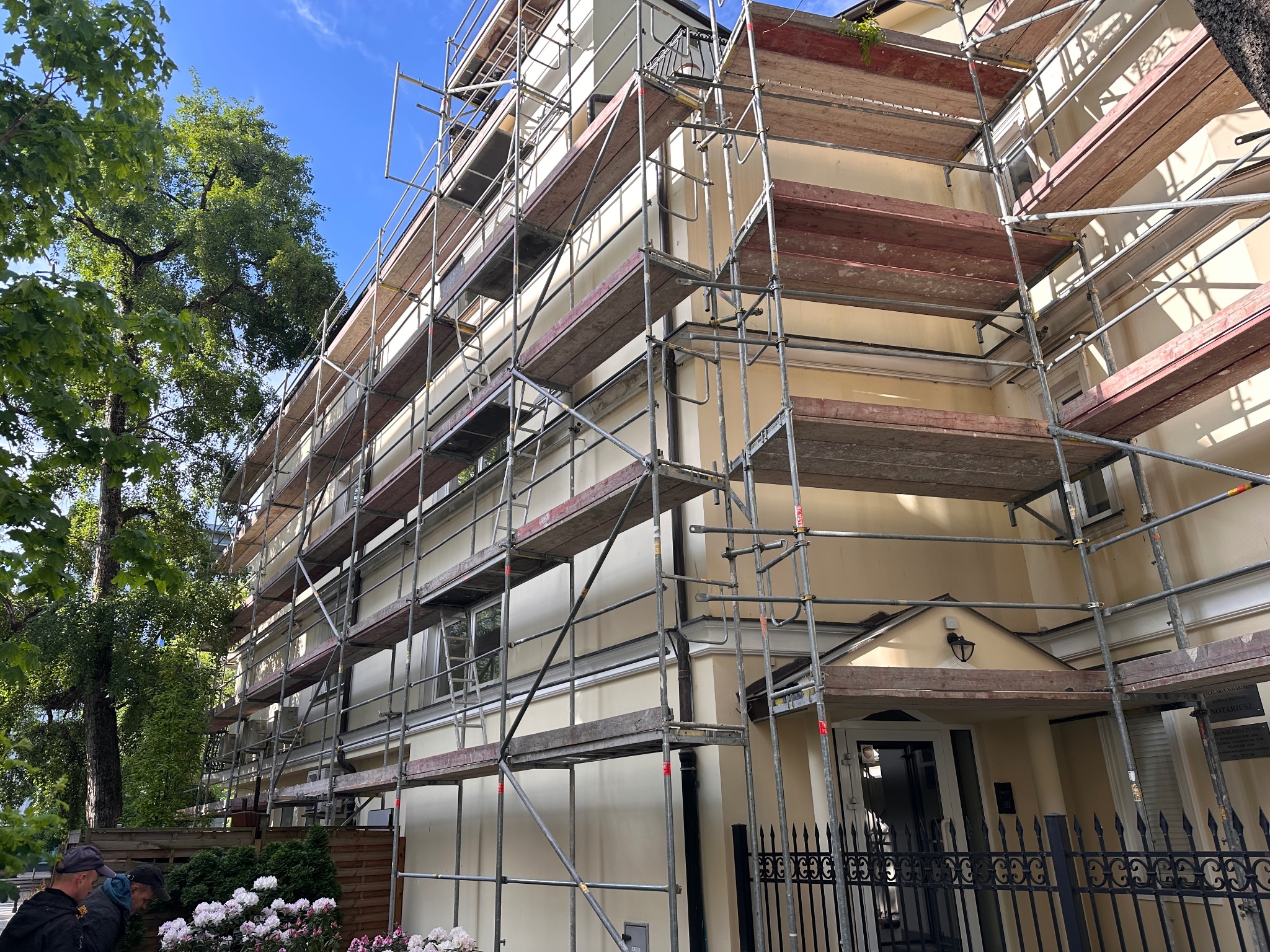 Elewacja budynku w trakcie prac. Widoczne rusztowania, fragment zieleni i ogrodzenie. Jasna kolorystyka budynku. Ujęcie z dołu, podkreślające wysokość.