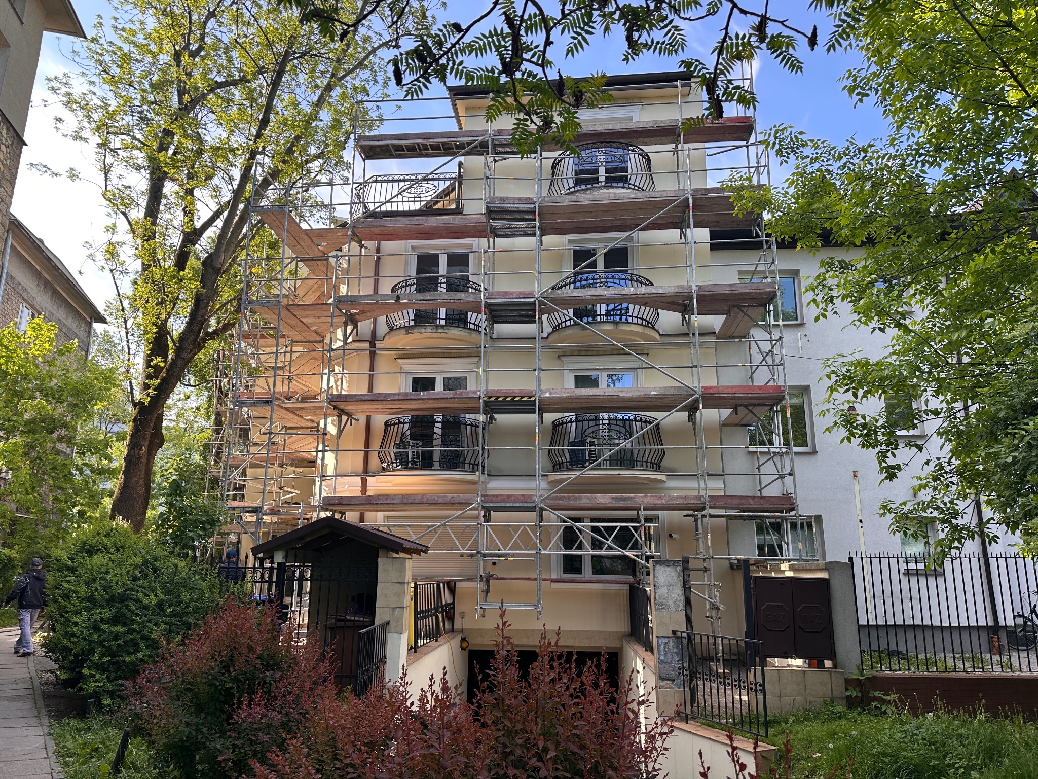 Budynek wielorodzinny w trakcie malowania elewacji, otoczony rusztowaniami. Widoczne balkony z ozdobnymi balustradami i zieleń drzew. Słoneczny dzień.