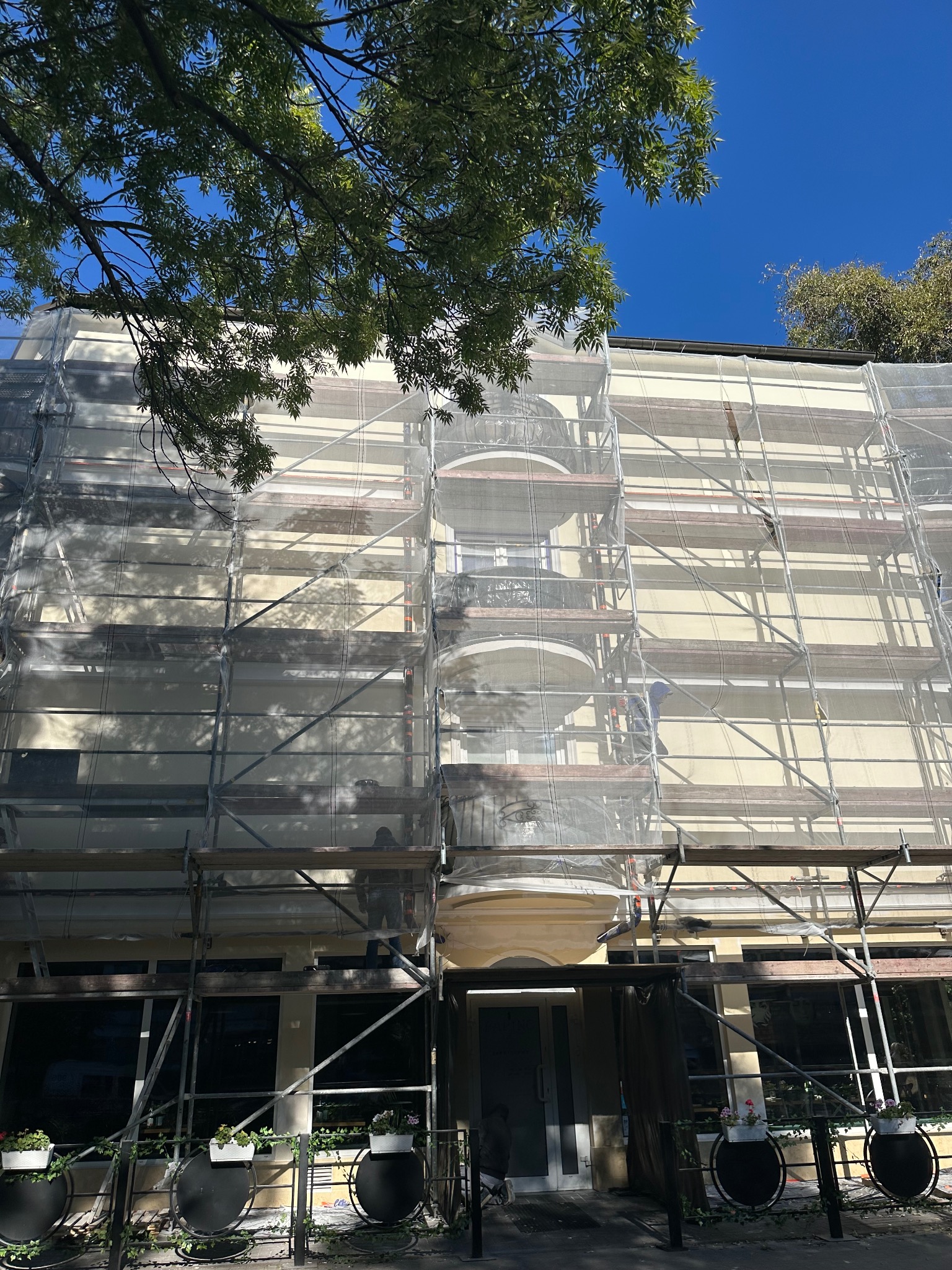 Budynek w rusztowaniach, zabezpieczony siatką, przygotowywany do prac na elewacji. Widoczne elementy dekoracyjne i zieleń u podnóża budynku.