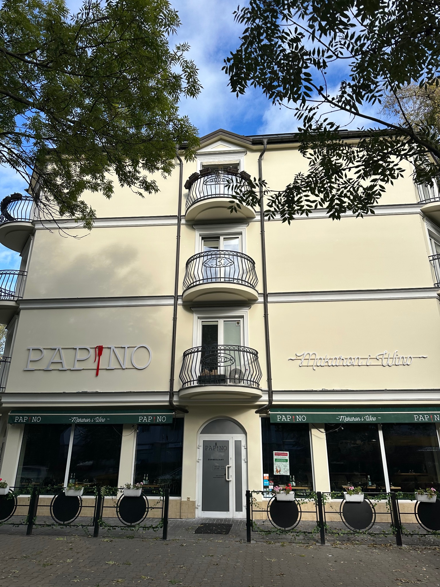 Elewacja budynku po odświeżeniu w Warszawie, z jasną farbą i dekoracyjnymi balkonami. Widoczne logo restauracji i zieleń drzew. Detal: czerwony element w logo.