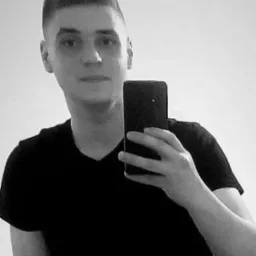 Czarno-białe selfie młodego mężczyzny trzymającego smartfon na tle jasnej ściany.