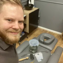 Autoportret mężczyzny z brodą w gabinecie w Śremie, na pierwszym planie leżanka z zagłówkiem, słoik z pijawkami, przybory do akupunktury i masażu, w tle umywalka i szafka.