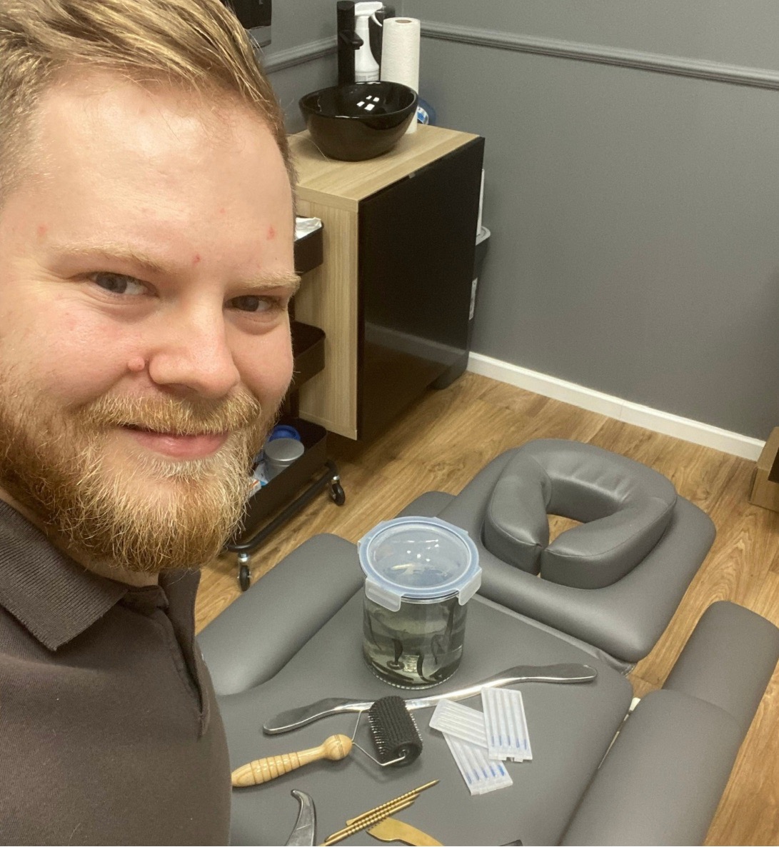 Autoportret mężczyzny z brodą w gabinecie w Śremie, na pierwszym planie leżanka z zagłówkiem, słoik z pijawkami, przybory do akupunktury i masażu, w tle umywalka i szafka.