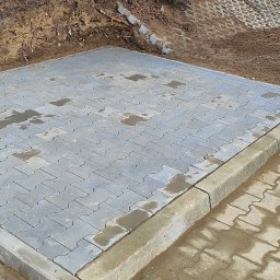 Eco-Dra-Bud. Patryk Drab - Ułożona, szara kostka brukowa w kształcie litery I, obrzeża z kostki, fragment paczkomatu po lewej, ziemia i śmieci w tle. Część kostki jest mokra.