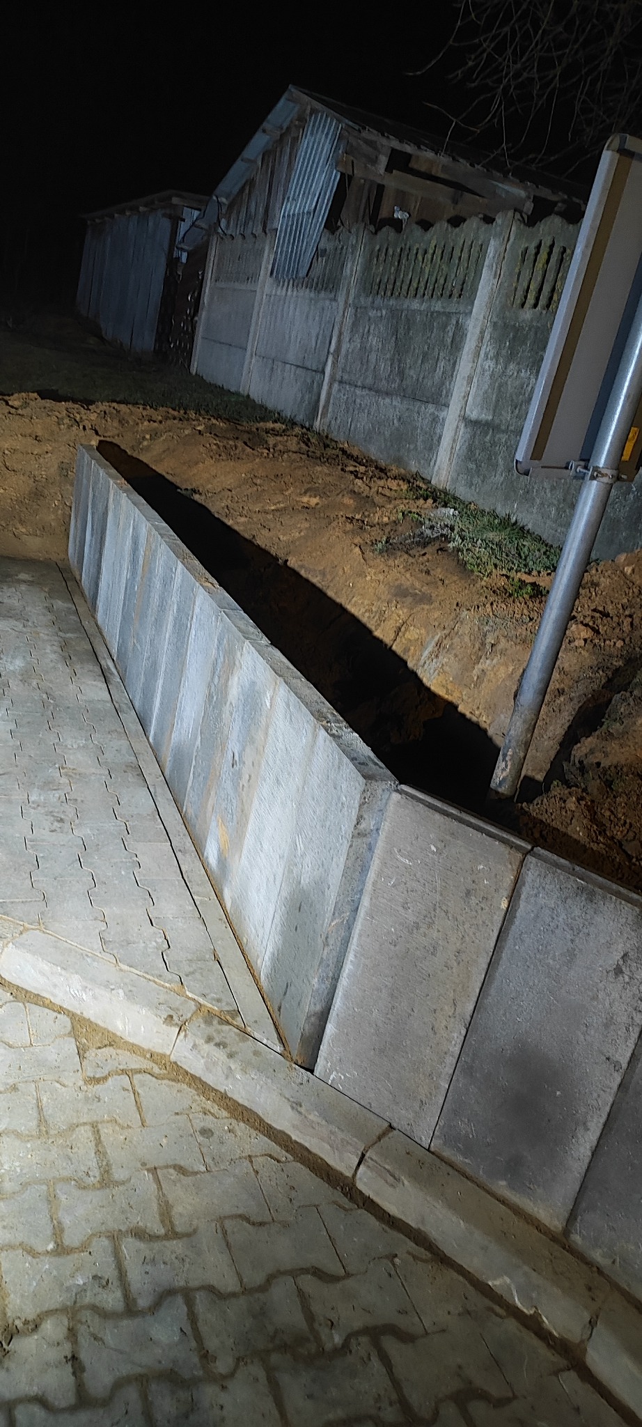 Ułożona kostka brukowa z obrzeżami i betonowym murem oporowym wzdłuż wykopu, oświetlona nocą. W tle widoczny płot i budynek gospodarczy.