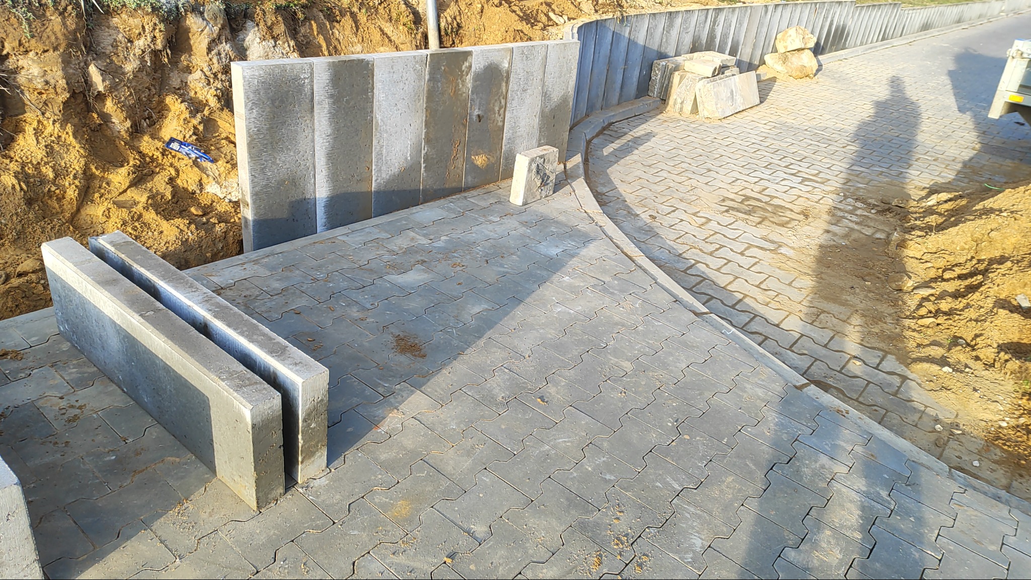 Ułożona szara kostka brukowa z obrzeżami, fragment wykopu ziemnego i betonowy murek oporowy. Widoczny cień osoby i pojazdu. Częściowo ukończona realizacja.