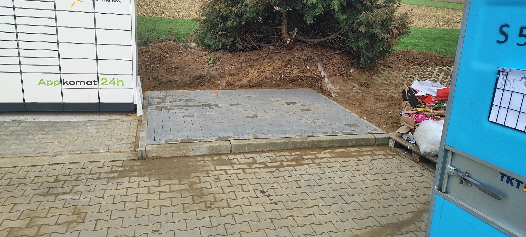 Ułożona kostka brukowa przy paczkomacie i przygotowany teren pod nasadzenia. Widoczne ślady po deszczu na kostce. Częściowo widoczny kontener na śmieci.