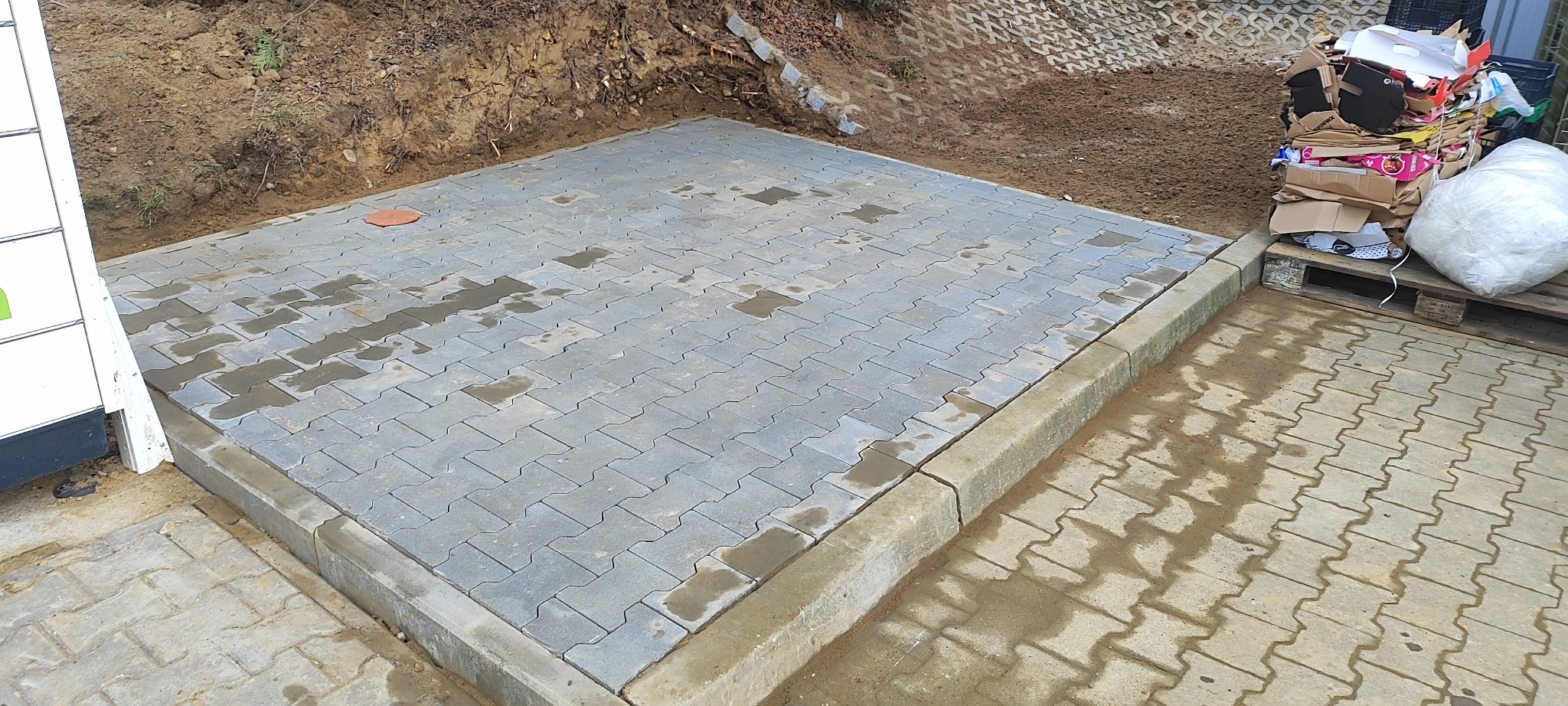 Ułożona, szara kostka brukowa w kształcie litery I, obrzeża z kostki, fragment paczkomatu po lewej, ziemia i śmieci w tle. Część kostki jest mokra.