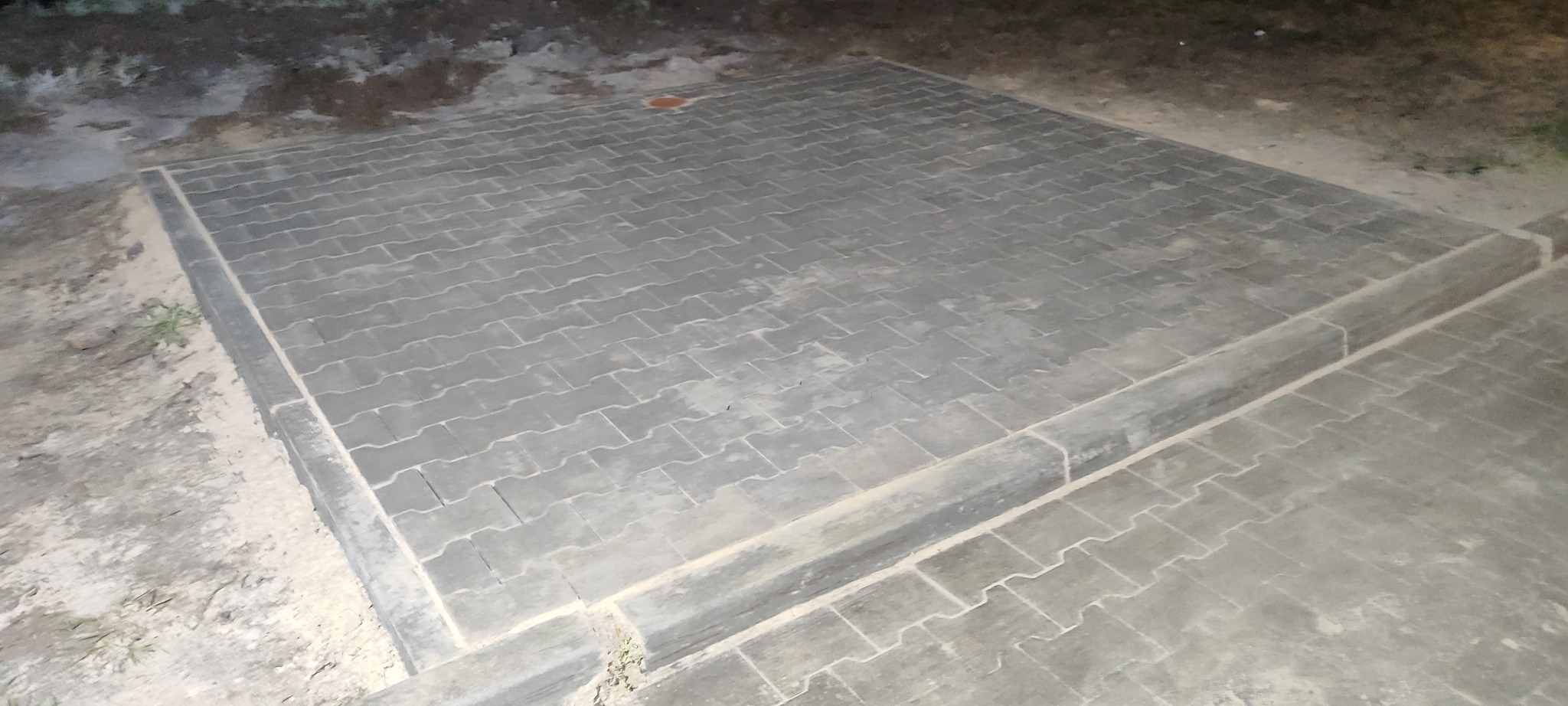Ułożona szara kostka brukowa tworząca kwadratową powierzchnię, obrzeża wykończone kostką krawężnikową. Widoczne ślady piasku na powierzchni kostki.