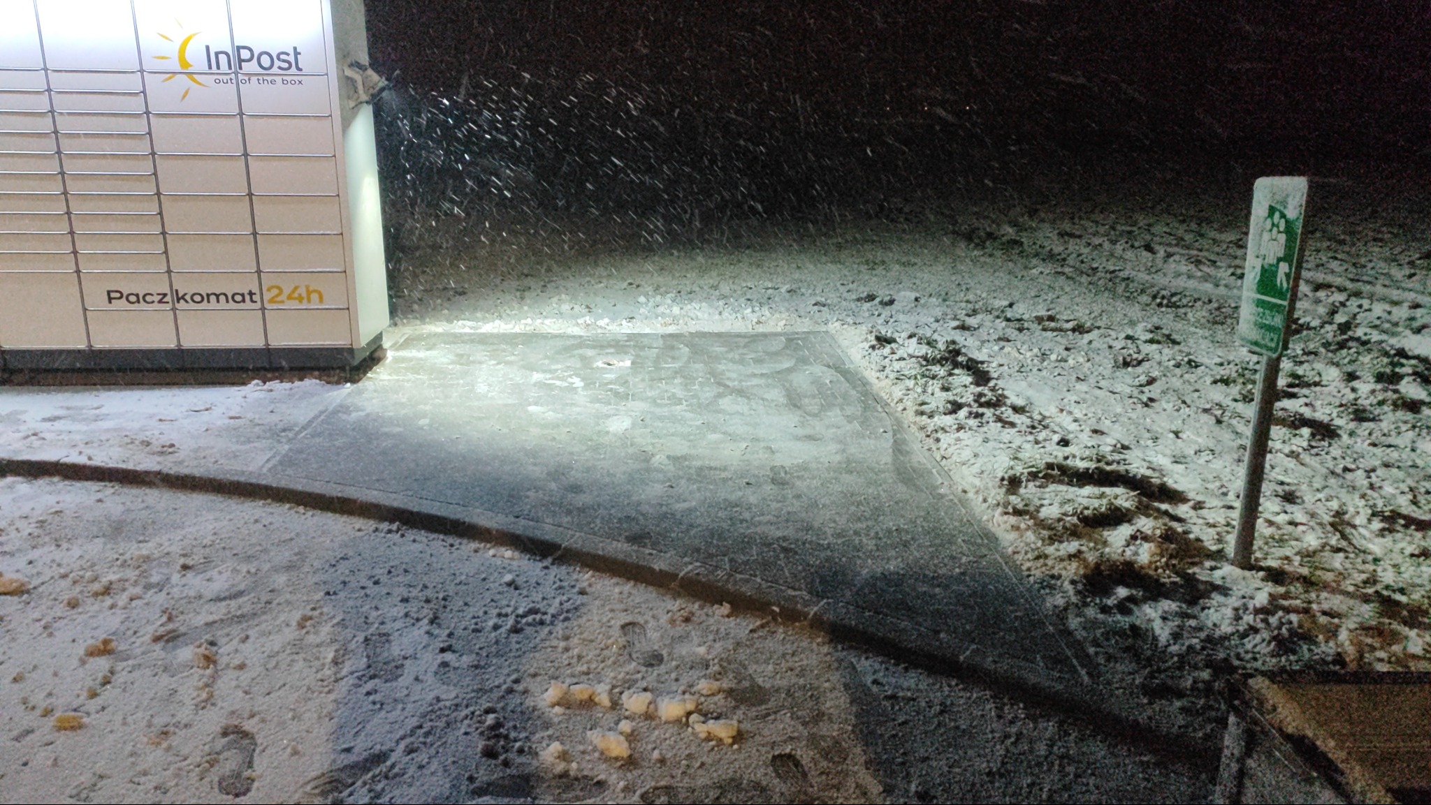 Zimowy widok na chodnik z kostki brukowej przy paczkomacie InPost w Wieluniu, pokryty śniegiem. Widoczny znak drogowy i padający śnieg.