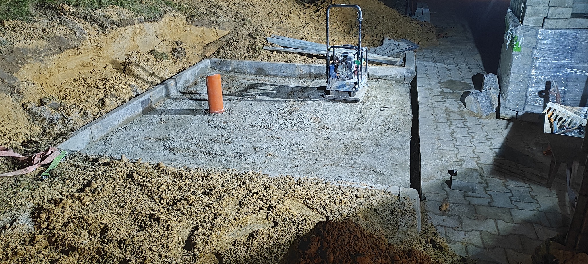 Przygotowany wykop z obrzeżami, zagęszczarką i rurą drenażową. Częściowo ułożona kostka brukowa w tle. Widoczne narzędzia i materiały budowlane.