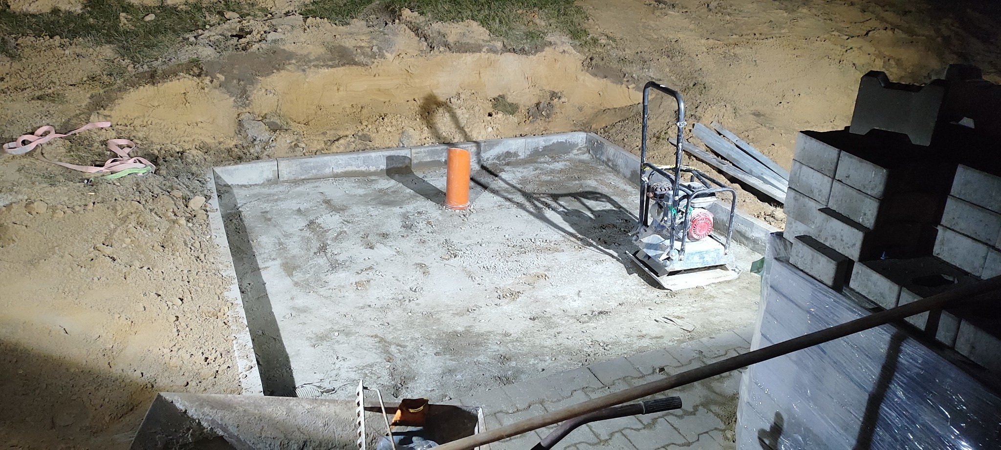 Przygotowany teren pod kostkę brukową z obrzeżami, zagęszczarką i rurą kanalizacyjną. Częściowo ułożona kostka brukowa w tle. Widoczne narzędzia i materiały budowlane.