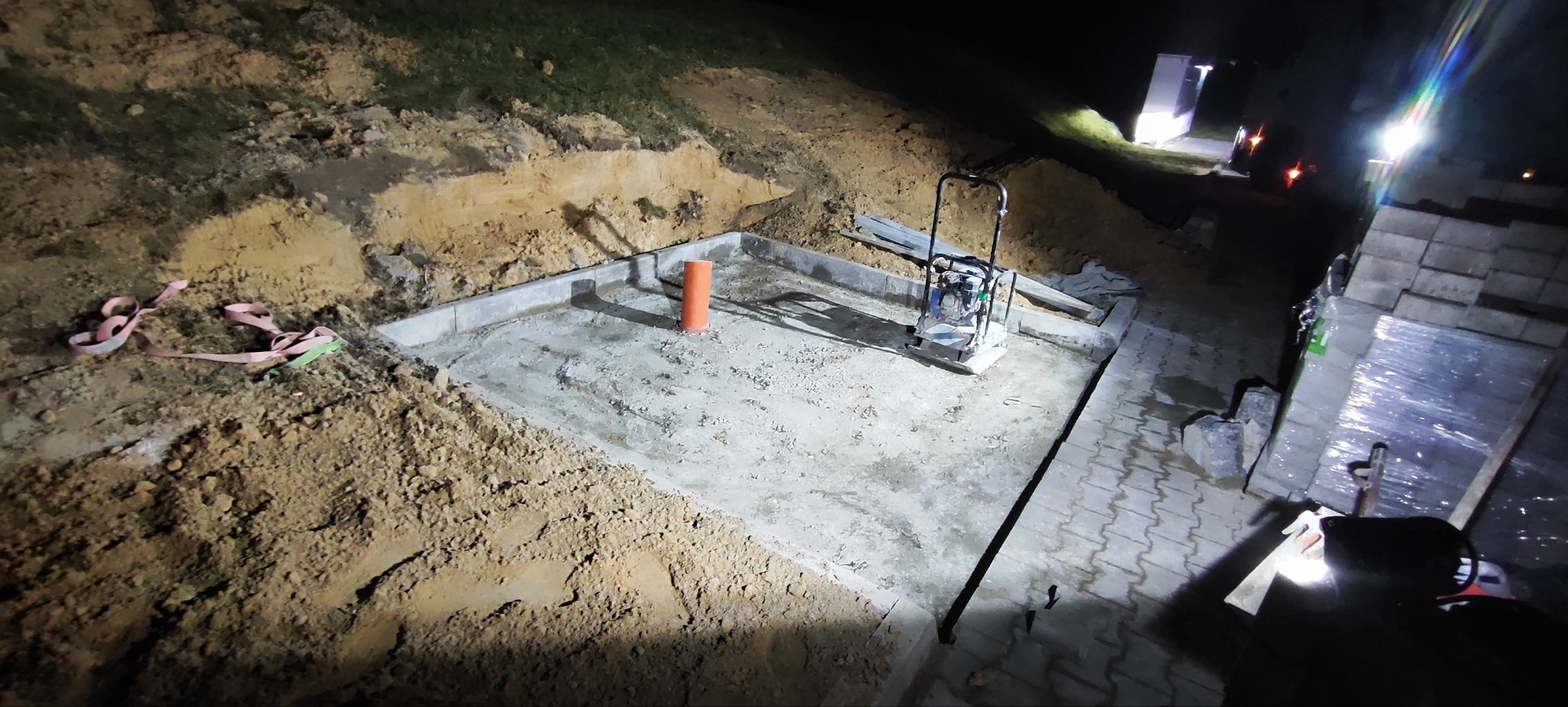 Nocne utwardzanie podłoża pod budowę: ubijarka wibrująca na betonowej płycie, obok stos kostki brukowej i wykop z ziemią. Pomarańczowa rura wentylacyjna.