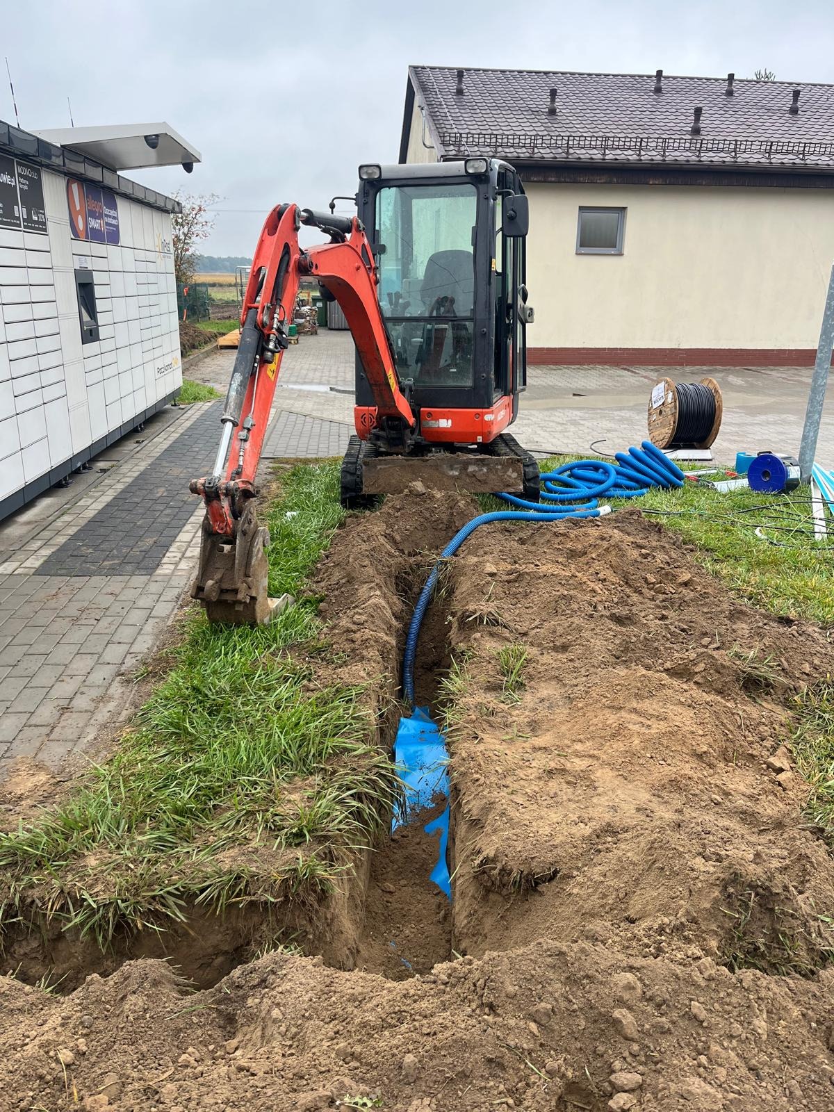 Mini koparka obok wykopu z niebieską rurą w ziemi, zabezpieczoną folią. W tle budynek i paczkomat. Widoczne kable na szpuli.