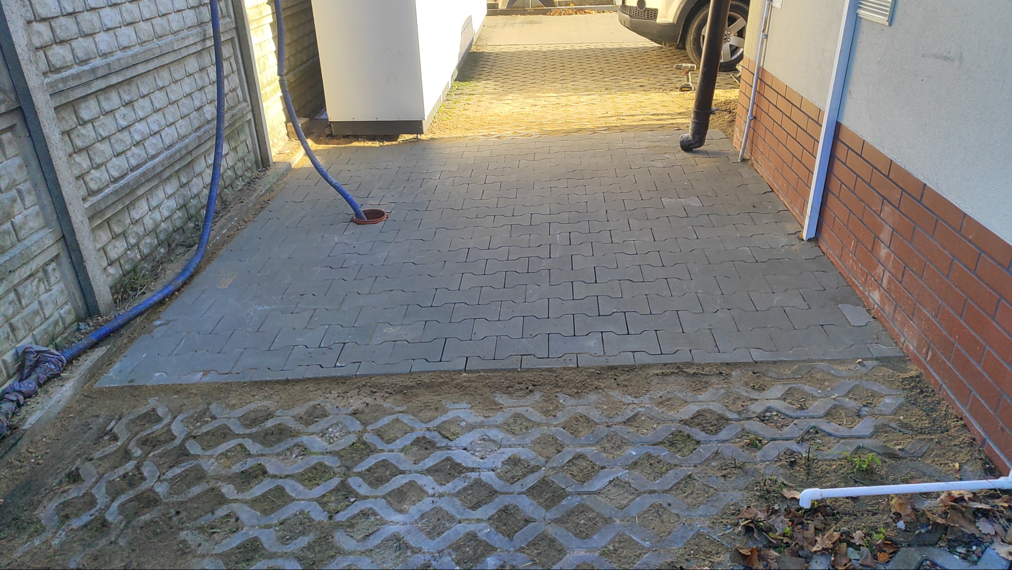 Ułożona szara kostka brukowa na posesji, widoczne obrzeża z ażurowych płyt. W tle budynek z ceglaną elewacją i fragment zaparkowanego samochodu.