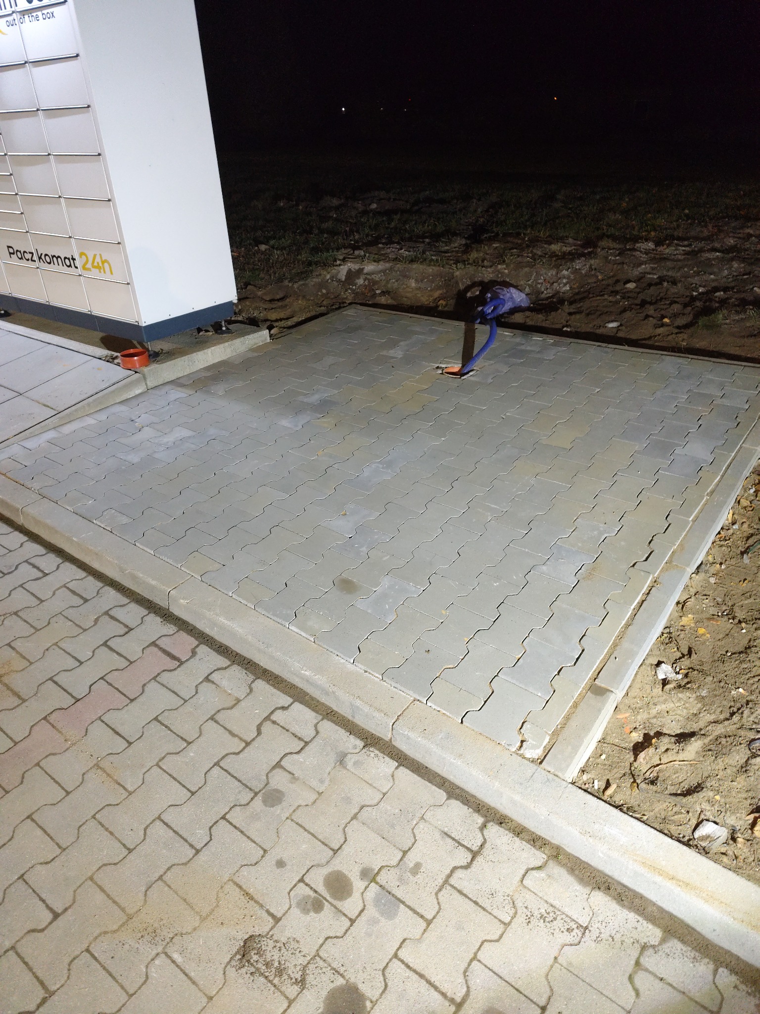 Ułożona kostka brukowa przy Paczkomacie, obrzeża betonowe, nocne oświetlenie. Widoczny fragment instalacji wystający z ziemi.