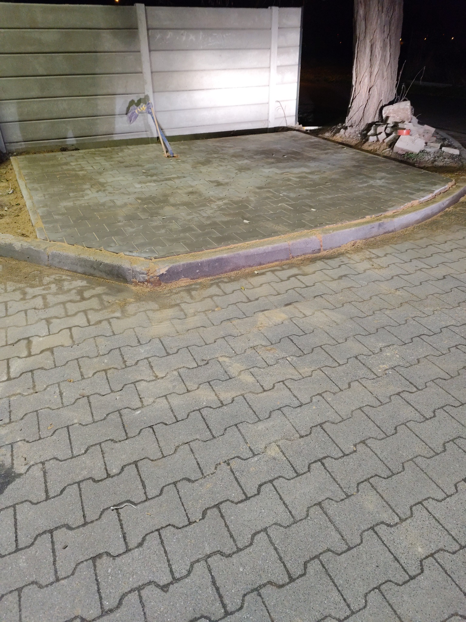 Ułożona szara kostka brukowa z obrzeżem, widoczna łopata oparta o betonowy płot. Część kostki jest mokra, a na obrzeżu widać piasek.