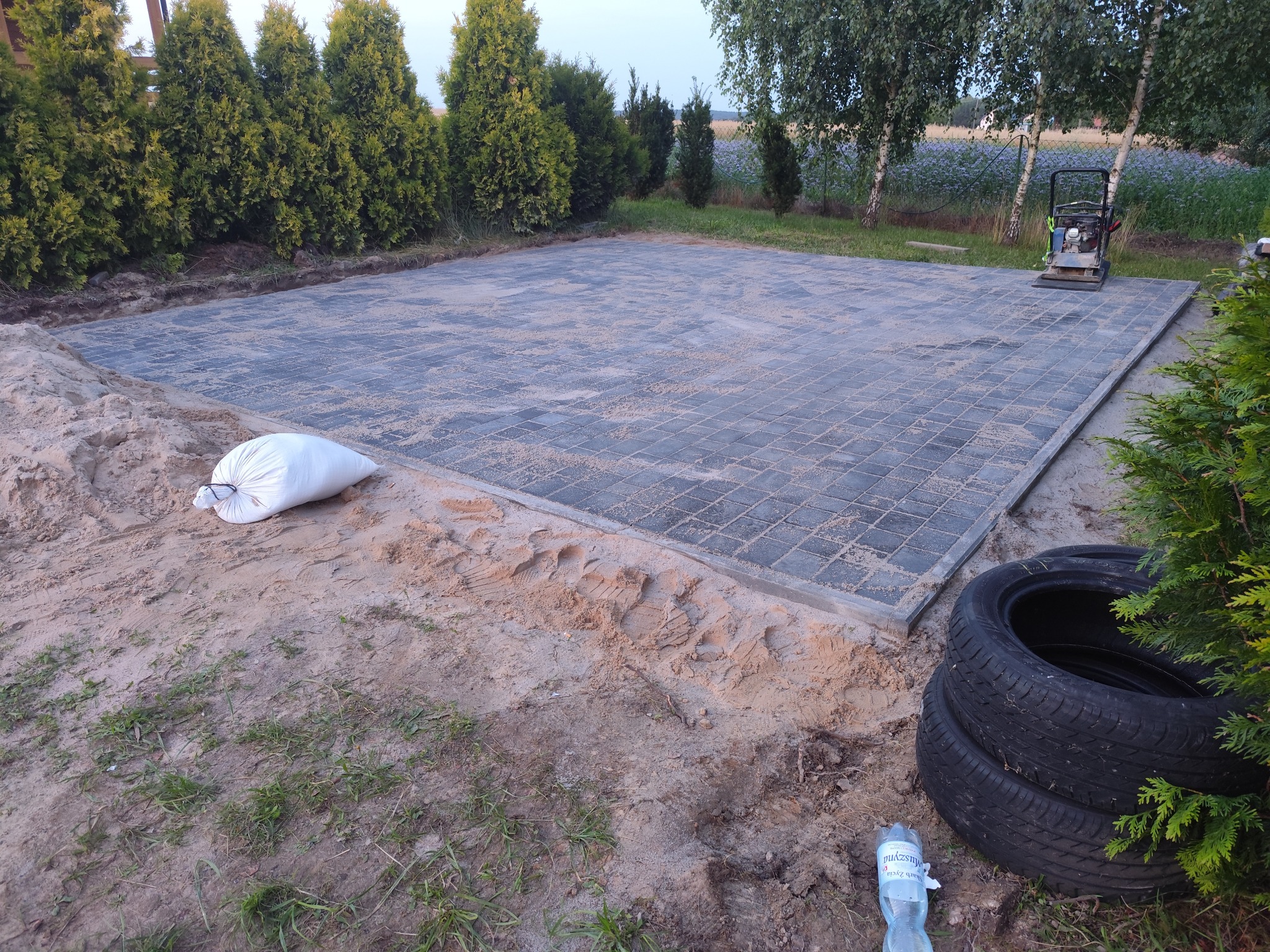 Ułożona szara kostka brukowa na podsypce piaskowej, z ubijarką w tle, otoczona zielenią i workiem piasku. Widoczne opony i butelka wody. Kąt ujęcia z dołu.